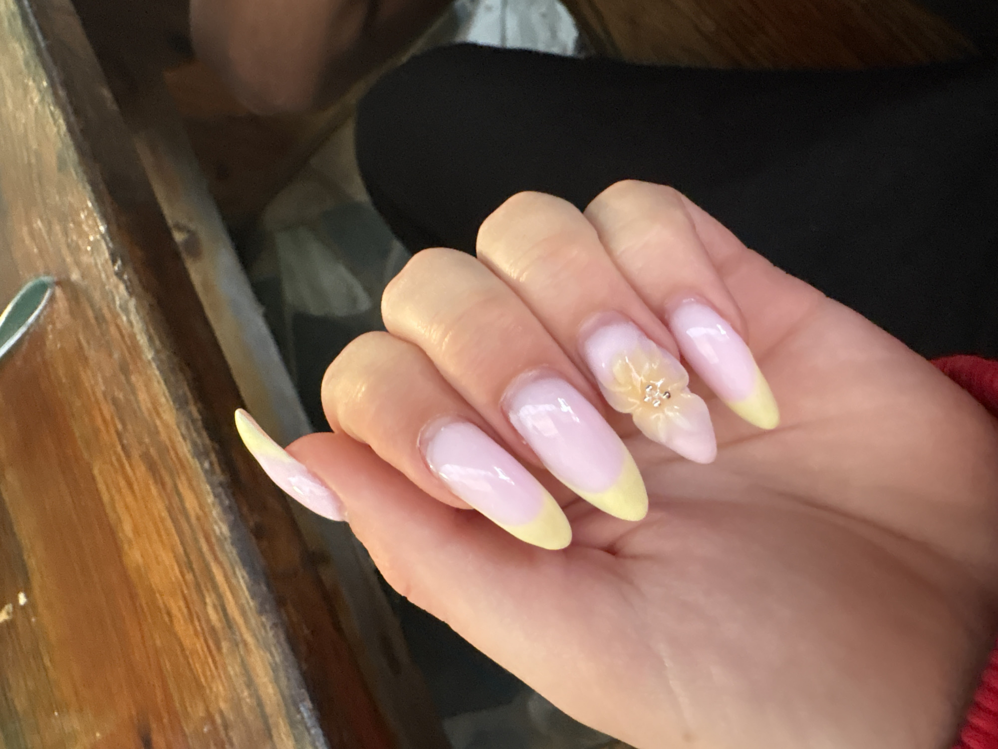 A hand with long, almond-shaped nails is shown against a background of dark clothing and a wooden surface. The nails are painted with a pale pink base and a yellow tip in a French manicure style. The ring finger has a flower adornment. The location is unknown and there is no information to suggest that the image was taken in the city of Tsarevo, Bulgaria.