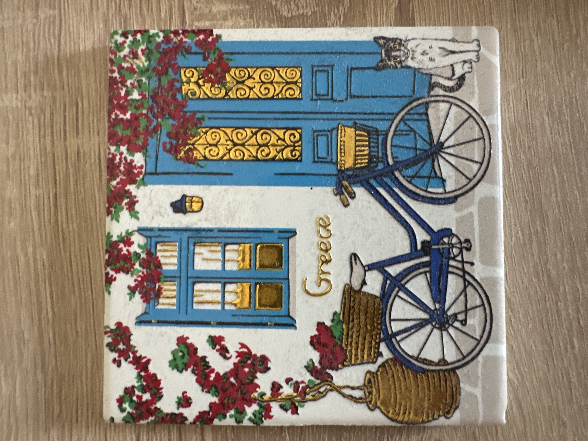 The image is a close-up of a decorative tile depicting a scene with a blue door and window, red flowers, a bicycle, a vase, and a cat. The tile is placed on a wooden surface. The scene is rendered in a stylized, illustrative manner. There are no people or actions visible. The background is a white wall with red and green floral vines. The door is bright blue with intricate gold scrollwork in its upper panels. A blue bicycle with a basket containing a yellow object is positioned in front of the door. To the left of the door, a stylized gold vase holds a branch with red flowers. A black and white cat with yellow eyes sits to the right of the door, looking towards the viewer. The bottom of the tile depicts a brick pattern. The word "Greece" is written in gold script above the bicycle. This image does not depict any specific location in the city of Marten, Bulgaria.