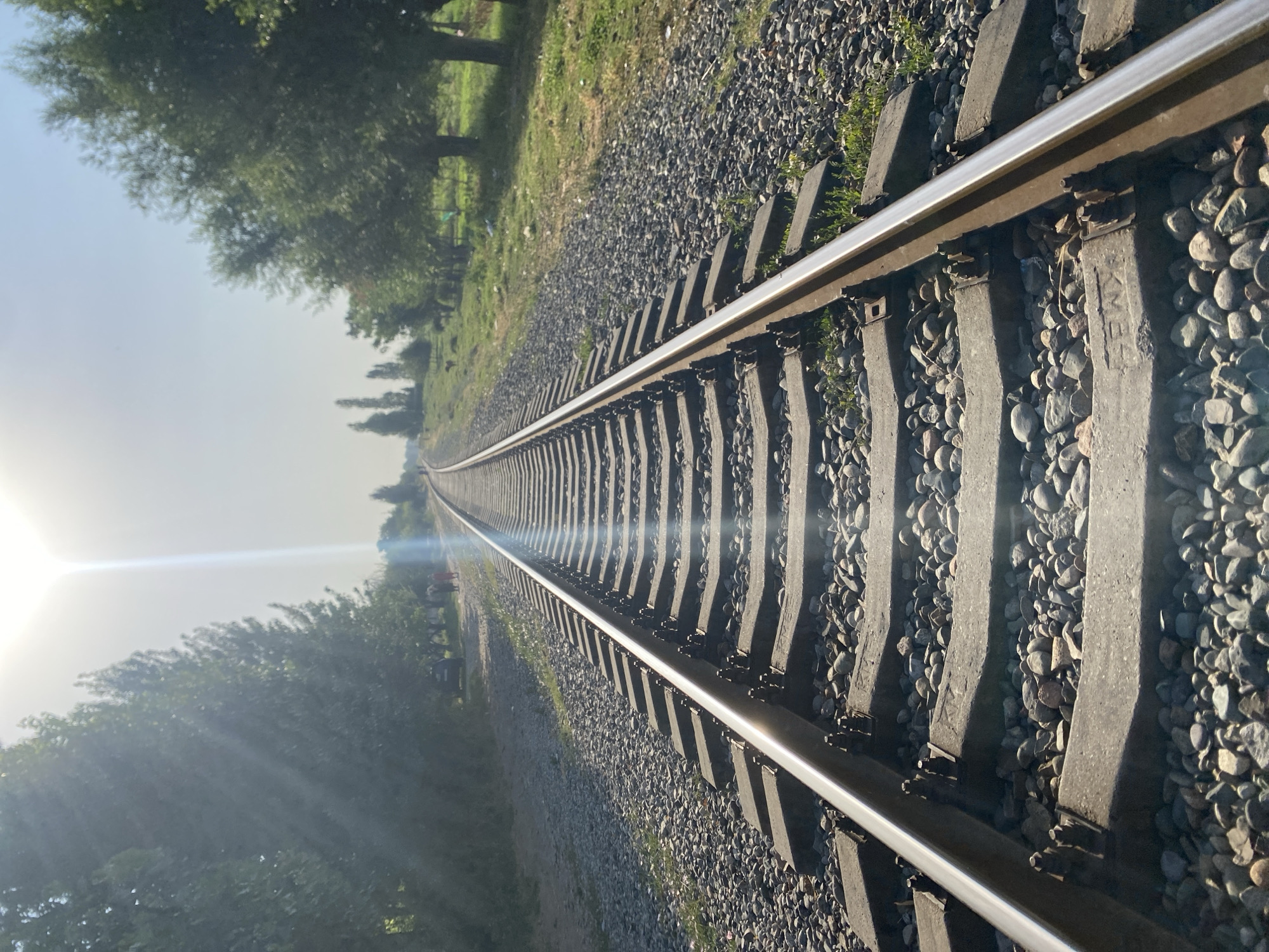 The image depicts a railway track in the city of Боғистон, Tajikistan. The perspective is low, looking down the length of the tracks, which recede into the distance. The tracks are made of metal rails supported by concrete sleepers, surrounded by gravel. On the left side of the tracks, there is a line of trees, and further back, a dark-colored car and two figures walking. On the right side, there are more trees and grassy areas. The sky is bright with the sun creating a lens flare and a visible light streak. The overall scene is illuminated by natural daylight, suggesting it is daytime.