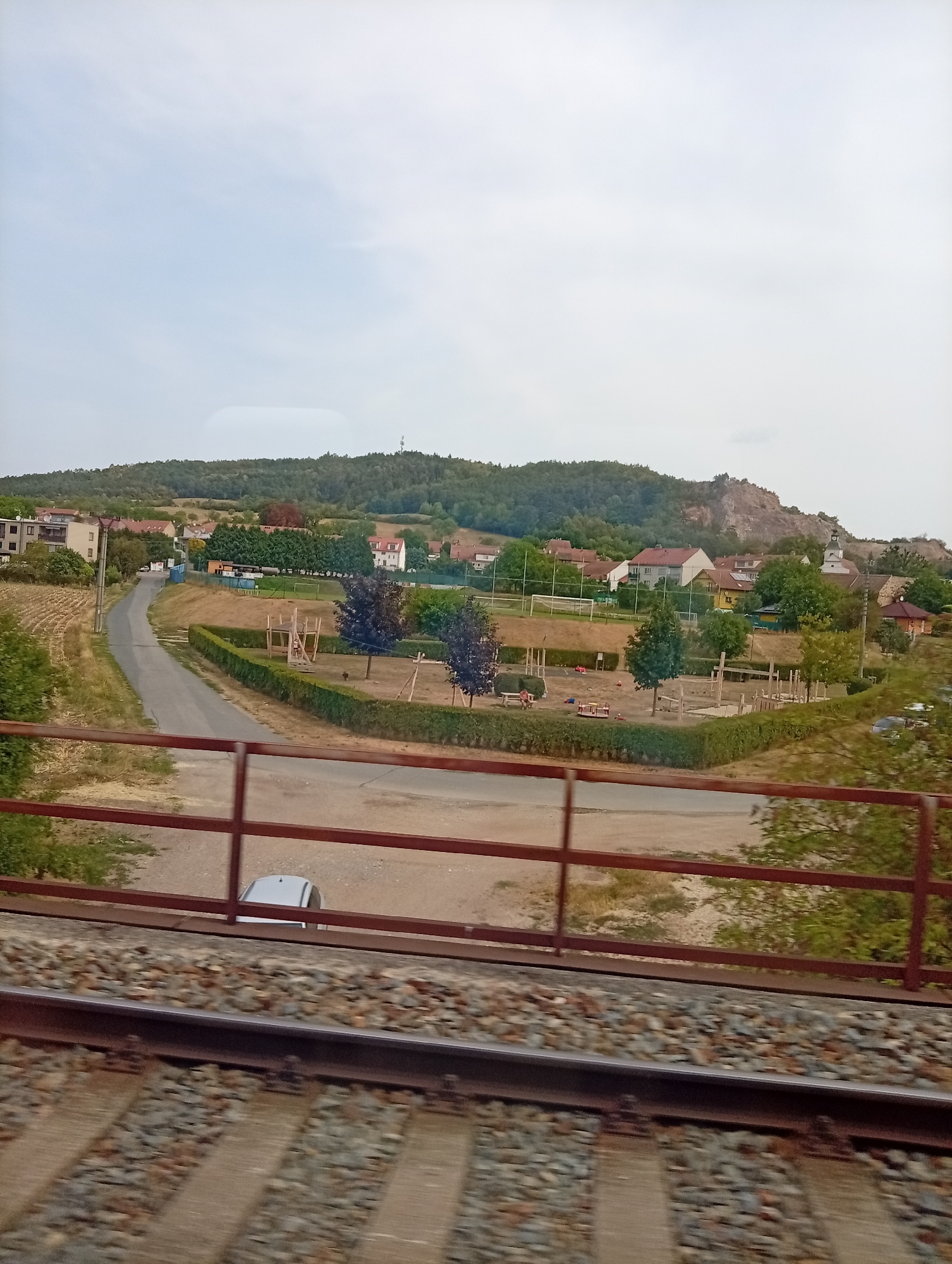 The image depicts a view from a train, looking out over a rural landscape that includes the town of Čebín, Czechia. In the foreground, train tracks with ballast are visible, partially obscured by a reddish-brown metal railing. Beyond the railing, a paved road curves through a grassy area. A playground with wooden structures and sandboxes is situated in the middle ground, bordered by hedges. Several trees are scattered throughout the scene. In the background, houses with red roofs form a village, nestled against a wooded hillside. A prominent rocky outcrop is visible on the hill. The sky is overcast. A telecommunications tower is visible on the distant hilltop.