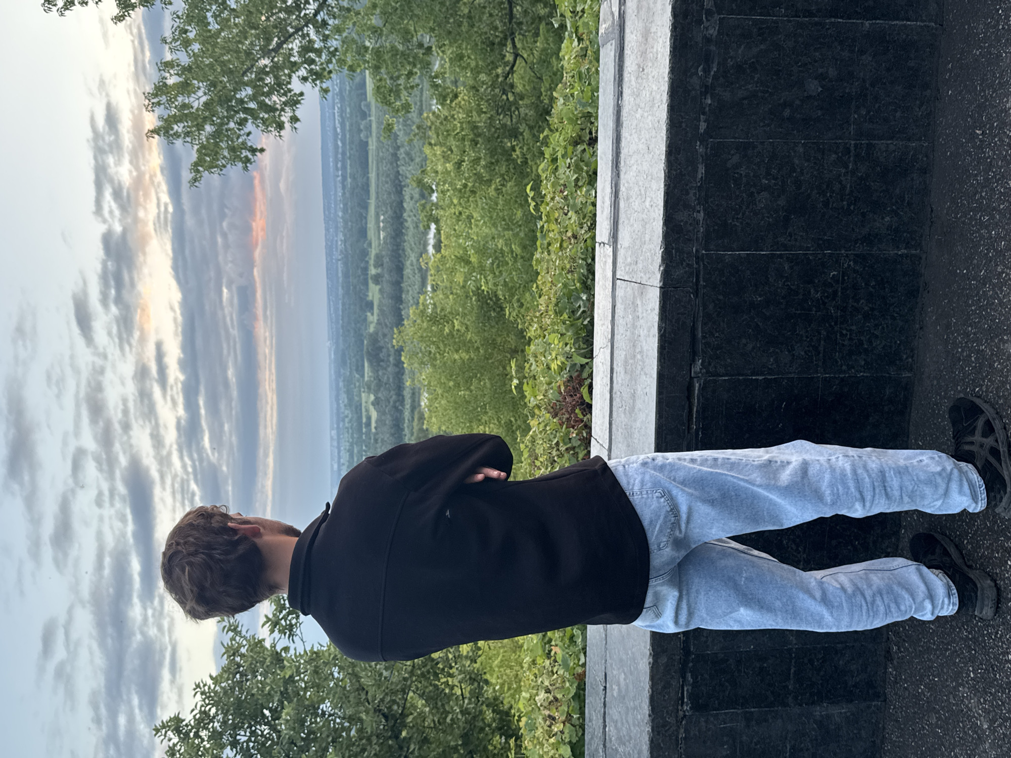An individual stands with their back to the camera, arms crossed, looking out over a landscape. They are wearing a black collared shirt and light-wash jeans, with black athletic shoes. The individual is positioned on a paved surface next to a dark stone wall. Beyond the wall, a lush green valley stretches out, dotted with trees and a river. The sky above is filled with dramatic clouds, with a hint of sunset colors near the horizon. The location is identified as the viewpoint near the monument to Salavat Yulaev in Ufa, Russia, offering a panoramic view of the Belaya River valley.