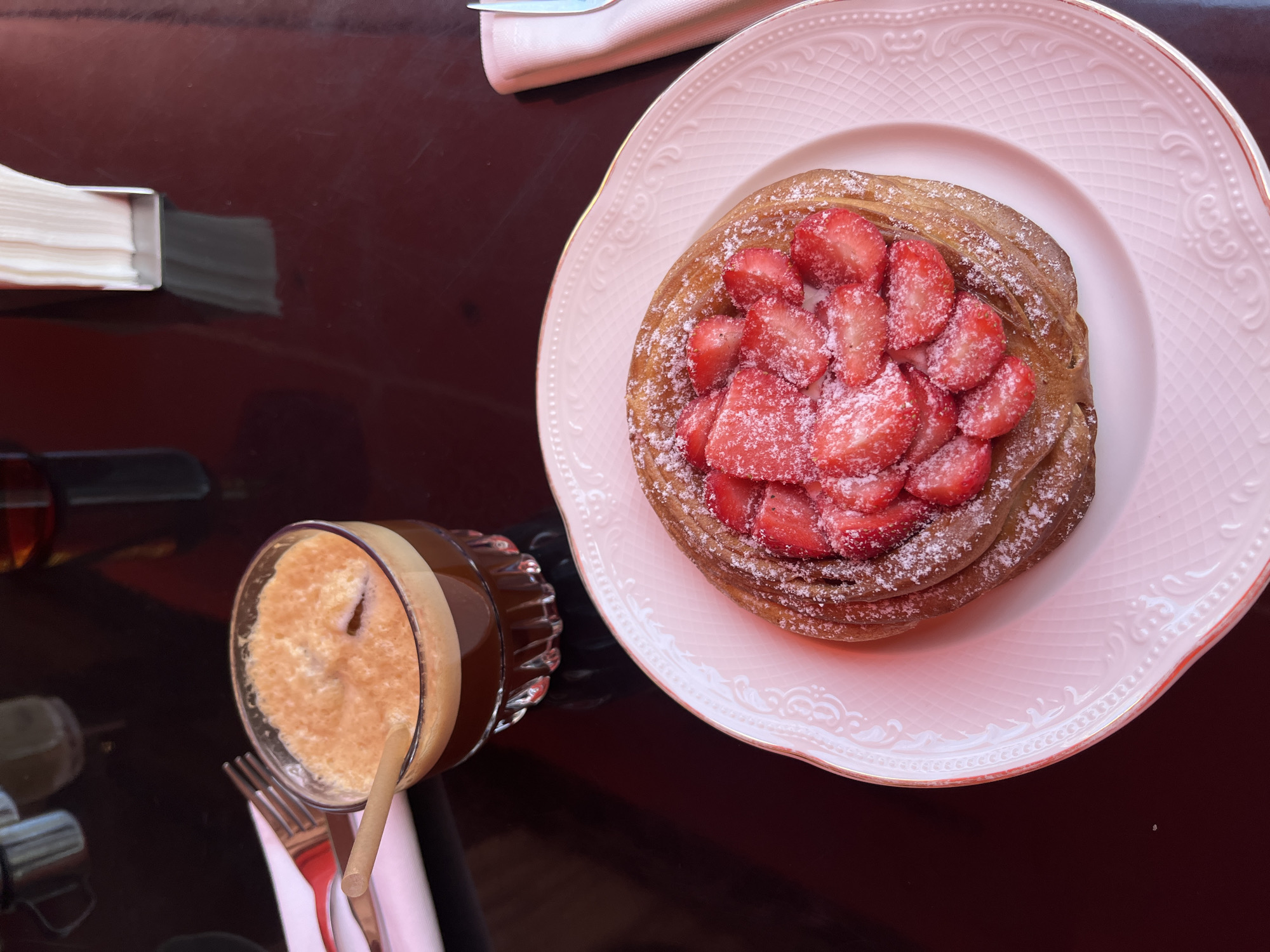 The image shows a table setting in a cafe, with a pastry topped with strawberries and powdered sugar served on a pink and white decorative plate. Next to it is a glass of iced coffee with a straw. Cutlery wrapped in a napkin is visible. A box of napkins and additional cutlery is also present. The glossy table reflects the items. The location is Yerevan, Armenia.