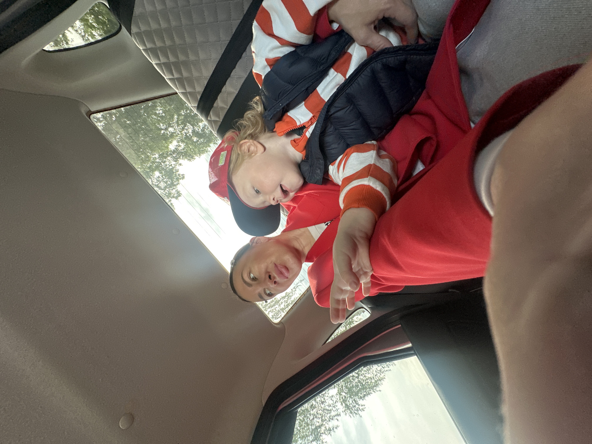 A woman in a red top is holding a young child in her lap inside a vehicle. The child wears a red and white striped shirt, a navy puffy vest, and a red baseball cap. A gray-quilted panel is visible in the car's interior, suggesting the roof area. The scene is oriented upwards, capturing the car's interior roof, windows, and portions of the outside environment, which appear to show trees under a cloudy sky. No clear indicators suggest Votkinsk, Russia as the location.