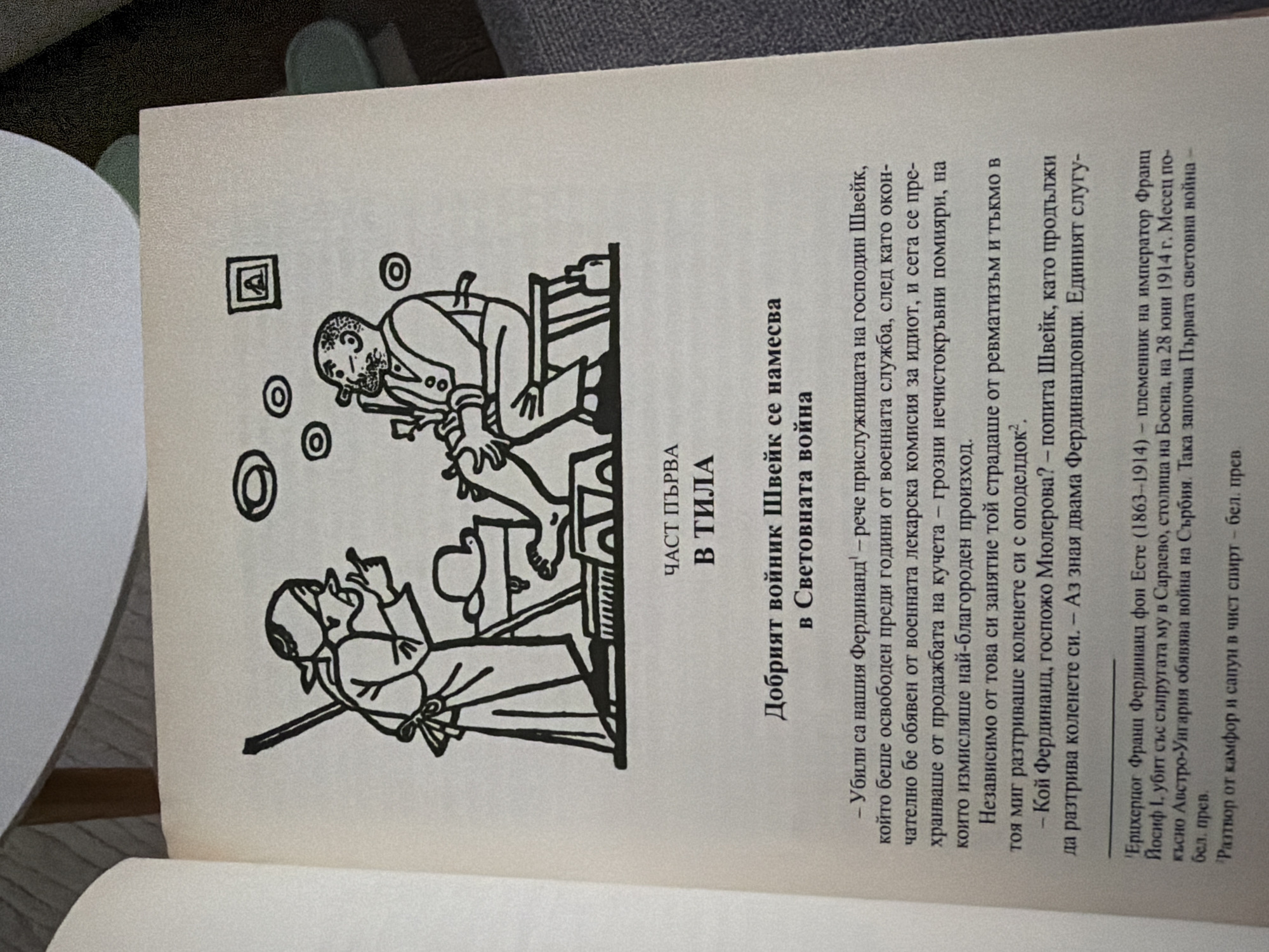The image is a close-up of an open book, displaying an illustration and text. The illustration depicts two figures in a cartoonish style: a woman with her finger raised in a scolding gesture, holding a broom, and a man seated, smoking a pipe. The man is wearing a vest and suspenders, and the woman is wearing an apron and headscarf. They are positioned near a table with some objects on it, including a hat. In the background, there are circular shapes and a framed picture on the wall.
Below the illustration, the book features text in Bulgarian. The heading reads "ЧАСТ ПЪРВА В ТИЛА" which translates to "PART ONE IN TILA". The following title is "Добрият войник Швейк се намесва в Световната война", meaning "The Good Soldier Švejk Intervenes in the World War". The text appears to be a narrative passage from the book, with dialogue and descriptive passages. There are also footnotes indicated by superscript numbers.
The question asks about the location in the city of Pazardzhik, Bulgaria. This information is not present in the provided image.