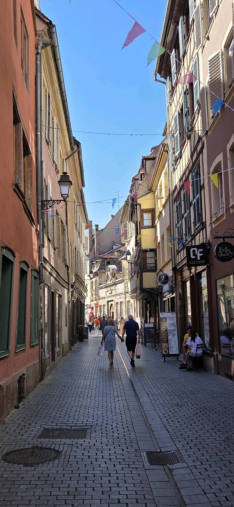 The image depicts a narrow cobblestone street in Strasbourg, France, lined with traditional Alsatian architecture. Buildings on either side feature timber framing, colorful facades (primarily orange and yellow), shutters, and steeply pitched roofs. Colorful triangular flags are strung across the street, creating a festive atmosphere.
In the foreground, a couple is walking away from the camera, holding hands. The woman wears a light blue dress and carries shopping bags, while the man is dressed in a dark shirt and shorts and also carries a shopping bag. Further down the street, other pedestrians are visible, some walking and some seated at outdoor cafe tables.
On the right side of the street, there are shops with awnings and signage. One sign reads "OF OZE FOOD" and another "Pasta Gusto". A menu board stands outside one establishment. A few people are seated at a small outdoor table in front of a shop.
The cobblestone pavement is well-maintained, with a distinct drainage channel running down the center of the street. A traditional street lamp is mounted on the left-hand building. The sky is clear and bright blue. The scene suggests a pleasant day in a charming European city.