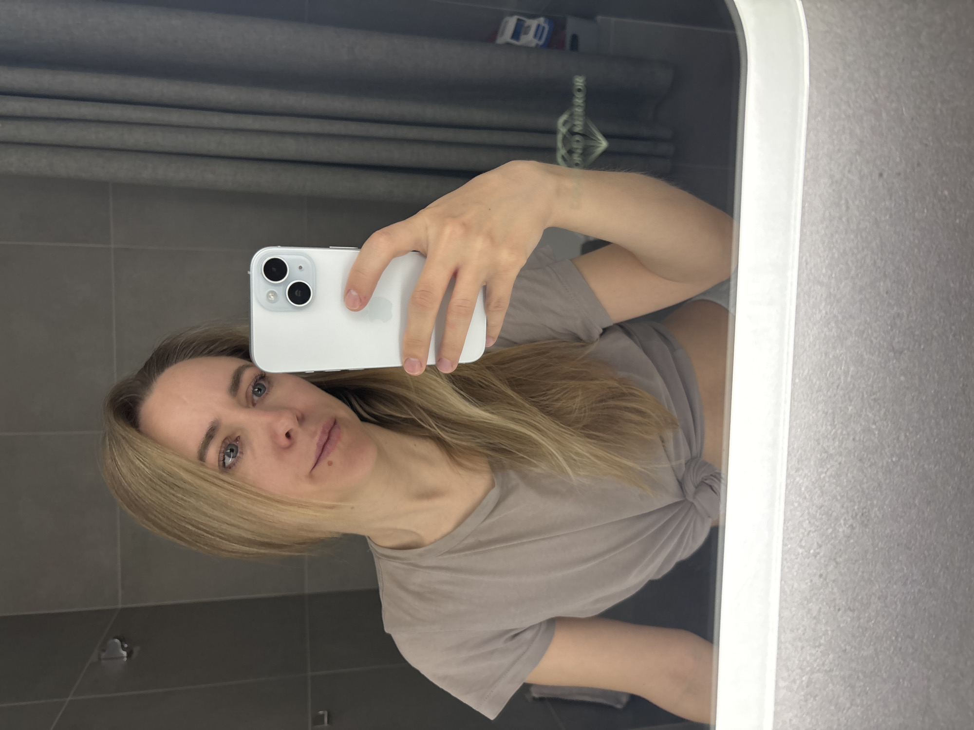 A woman is taking a selfie in a bathroom. She holds a white smartphone in her right hand, partially obscuring her face. She has fair skin, blonde hair, and blue eyes. She is wearing a taupe-colored t-shirt that is knotted on her right side. The bathroom appears to have gray tile walls and a mirror. The environment suggests a modern or recently renovated space. There is no information to indicate the photo was taken in Chaikovsky, Russia.