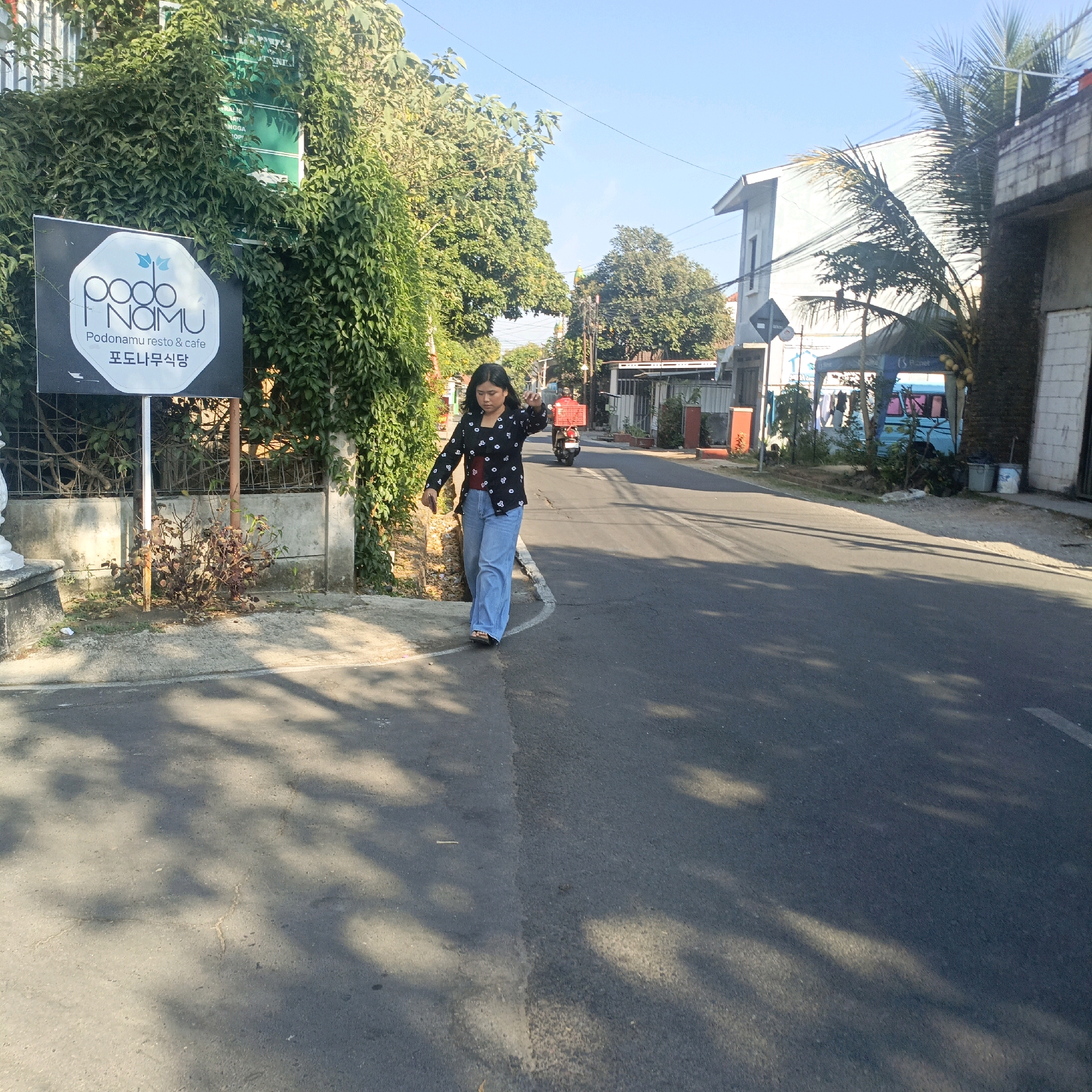 A young woman walks on the side of a paved road in Salatiga, Indonesia. She is wearing a black patterned shirt and light blue wide-leg pants, with her hair down. A sign for "Podo Nomu resto & cafe" is visible to her left, covered in lush green vines. The road is lined with trees and buildings, and a motorcycle with a red crate on the back is moving away from the viewer down the street. Sunlight casts shadows on the road, suggesting it is daytime.