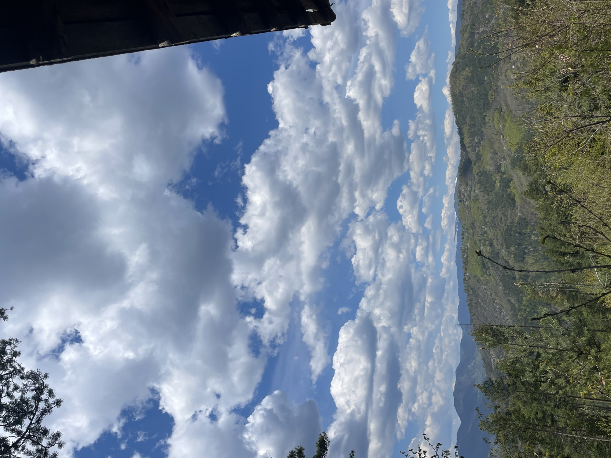 The image depicts a view towards a mountain range under a partly cloudy sky. The foreground includes parts of a building roof on the upper left and tree branches at the bottom. The sky is blue with white clouds. The mountain range has green vegetation. The location is assumed to be near Gradat, Bulgaria.