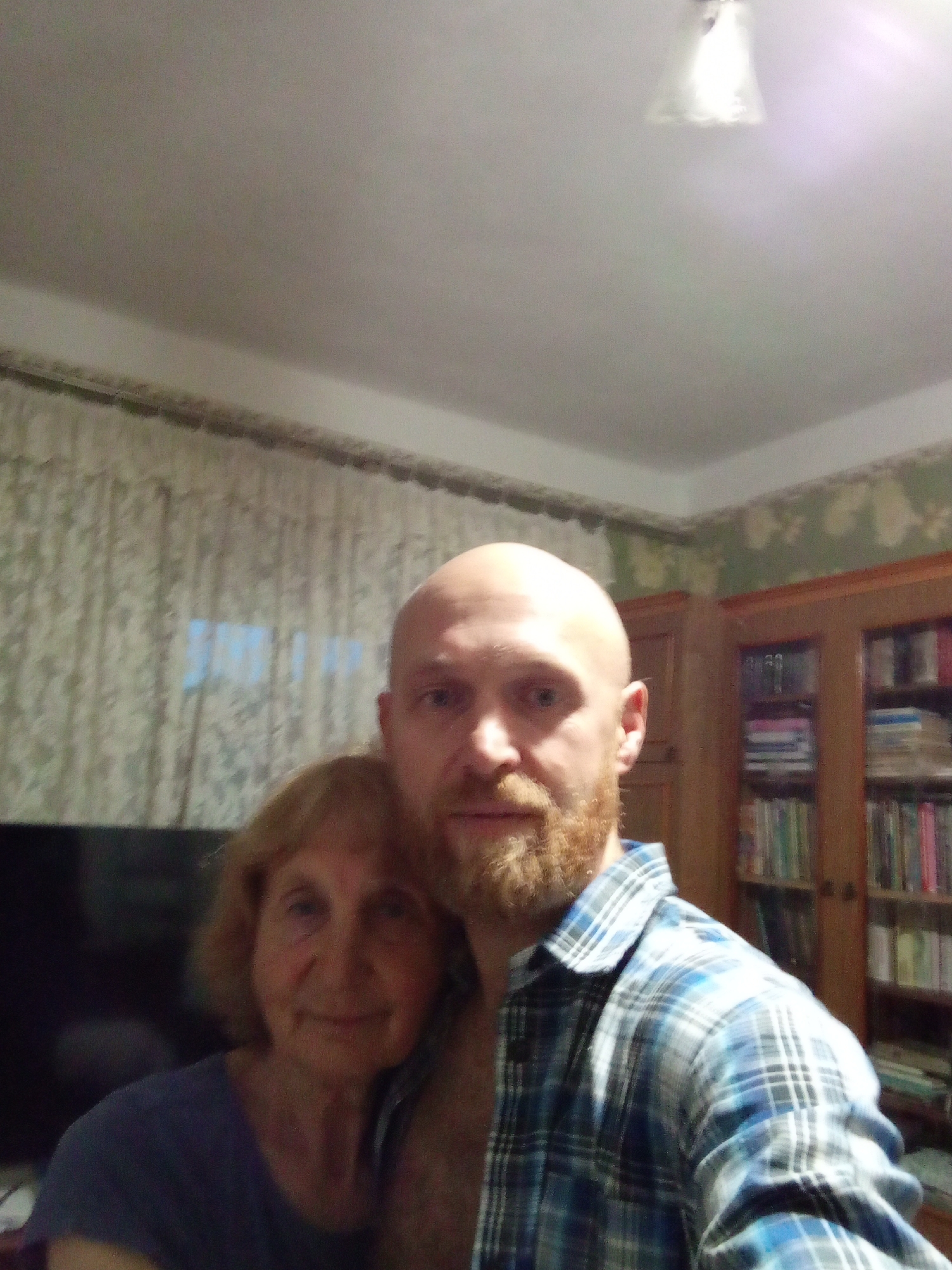 This media file is a selfie featuring two people in what appears to be a home setting. On the left is an older woman with blonde hair, wearing a dark purple shirt. She is smiling gently. To her right and slightly behind her is a bald man with a reddish-brown beard, wearing a blue and white plaid shirt. He is looking towards the camera.
The background shows a room with patterned curtains behind the woman, and to the right, a wooden bookshelf filled with books. The lighting suggests it might be evening or indoors, with a light fixture visible on the ceiling. The overall atmosphere seems relaxed and familial, with the two subjects posing together. There is no visible text in the image.