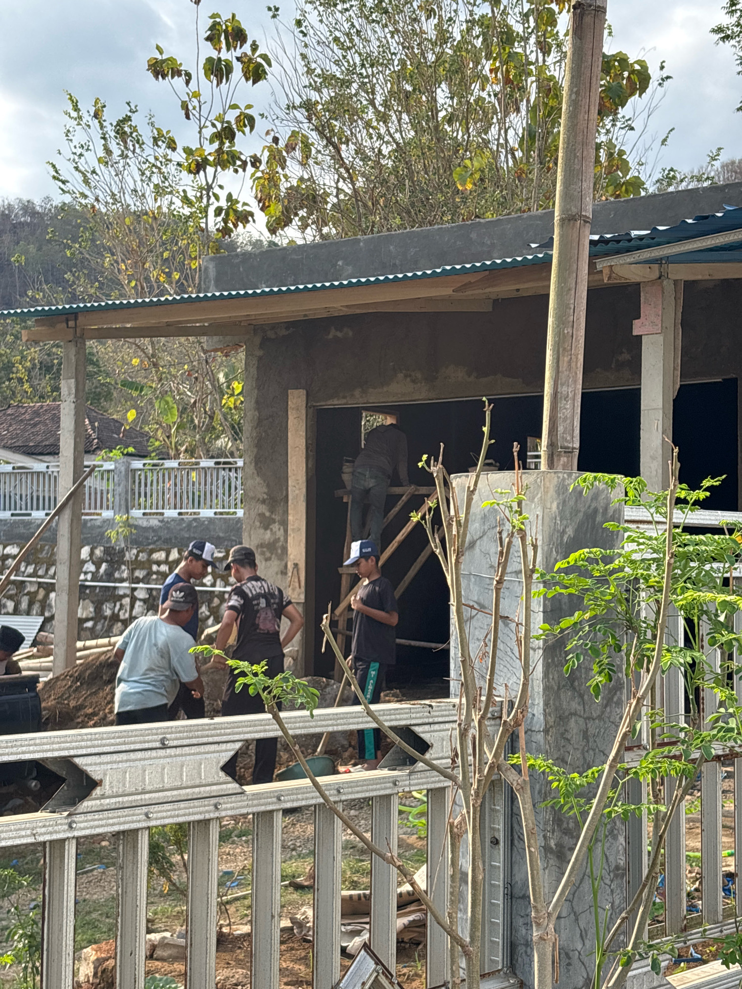 This image captures a construction scene in Tirtomoyo, Indonesia. Several individuals, appearing to be construction workers, are engaged in various activities related to building a structure. In the foreground, a man in a light blue t-shirt and a cap is focused on a pile of dirt or material. To his right, another worker in a dark t-shirt with white lettering and a cap is also looking down. In the background, a person in a black shirt and jeans is climbing a ladder to access the upper part of the building. Another young person wearing a blue t-shirt and a cap stands near a ladder. The structure itself appears to be in an early stage of construction, with concrete walls and a corrugated metal roof. The environment is outdoors, with trees and vegetation visible, suggesting a rural or semi-rural setting. The overall scene depicts ongoing construction work in a sunny, possibly humid climate.