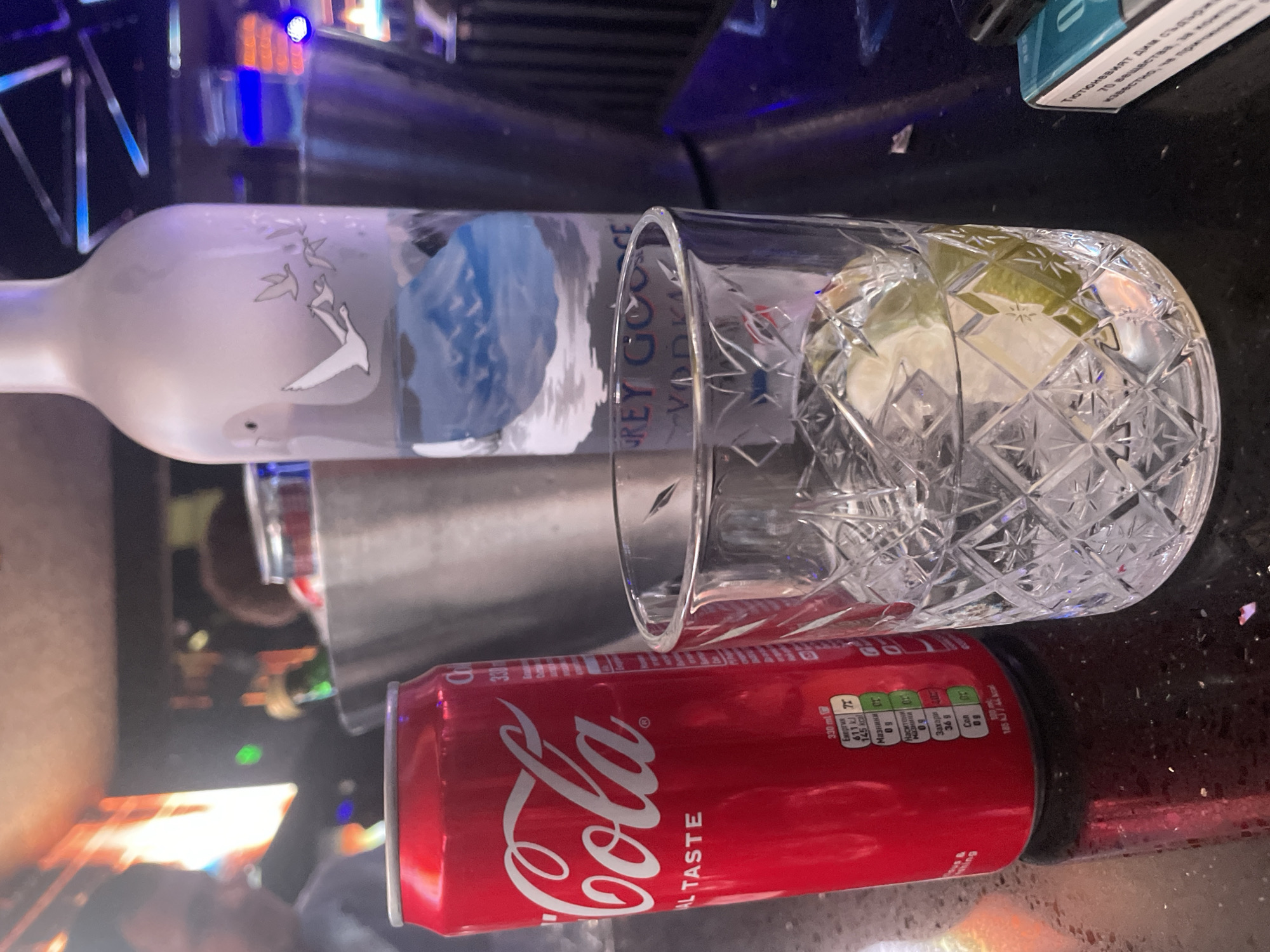 The image shows a Grey Goose vodka bottle lying horizontally on a dark, reflective surface alongside a crystal glass filled with ice and lime slices. Below the glass and vodka bottle is a red Coca-Cola can. The can is clearly labeled, including nutritional information in Bulgarian. The setting is dark, possibly a bar or home bar. The text on the Coca-Cola can indicates it is sold in Bulgaria, but there is no evidence to place the image in Sofia. A small portion of text at the top corner of the image contains Bulgarian script, possibly related to tobacco warnings, further confirming the Bulgarian connection.