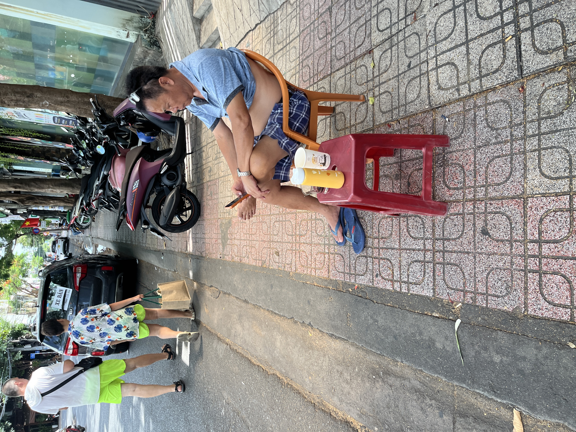 In a city street in Nha Trang, Vietnam, a man sits on an orange plastic chair on a tiled sidewalk. He is wearing a blue short-sleeved shirt and checkered shorts, with a pair of sunglasses perched on his head. He is holding a mobile phone in his right hand and appears to be looking at it. In front of him, on a red plastic stool, are two cups, one yellow and one white, and a glass. To the left of the man, two other people, possibly a father and son, are walking away from the camera. The man in front is wearing a white hooded t-shirt and bright green shorts, while the child is wearing a floral patterned shirt and green shorts, carrying a brown paper bag. Behind them, a dark-colored car with a "XE BÁN" sign is parked on the street. The street is lined with trees and various parked motorcycles. The overall scene depicts a typical urban environment in Vietnam on a sunny day.