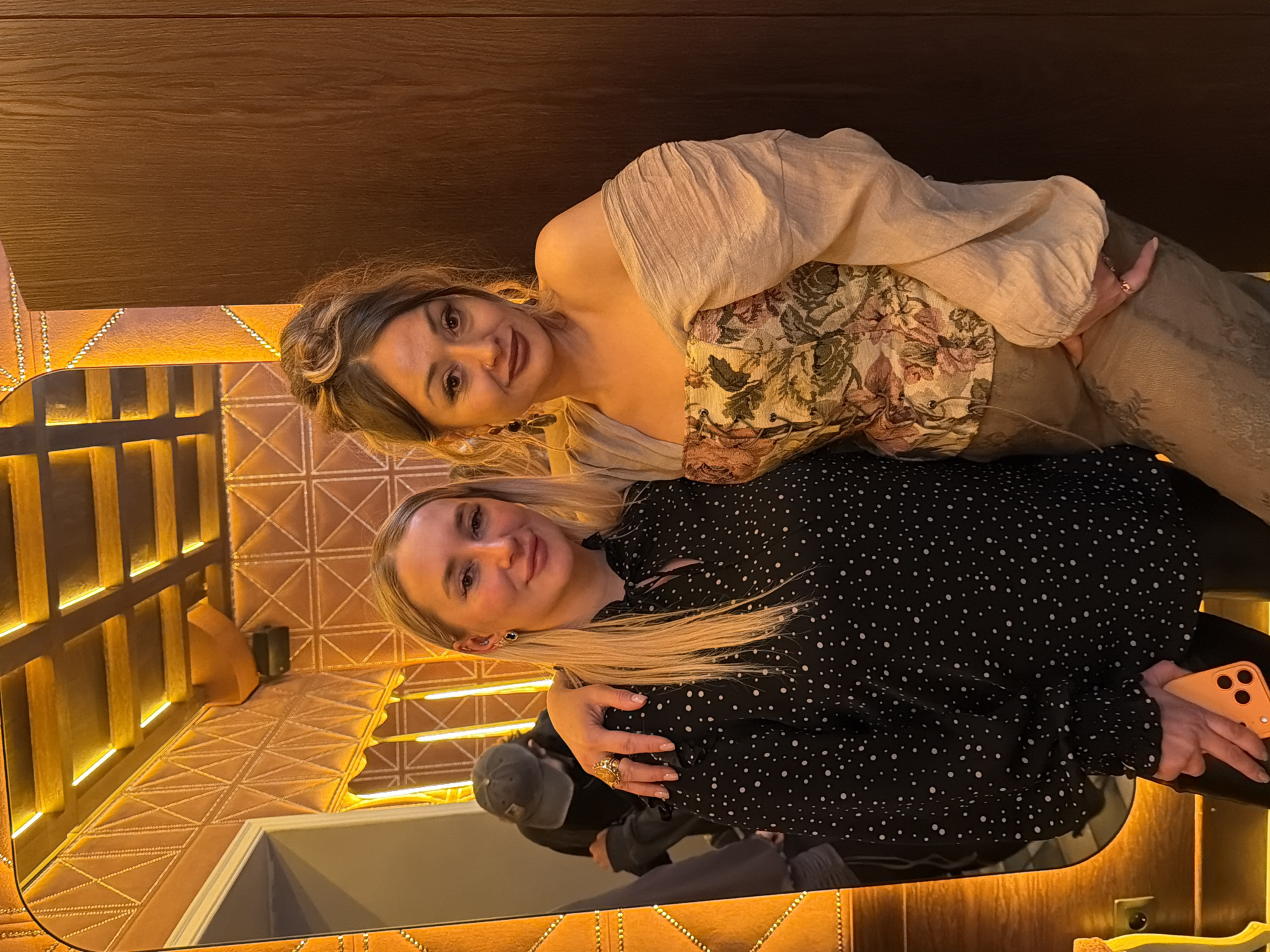 Two women are posing in a restroom with reflective decor, likely at a restaurant or bar in Krasnoyarsk, Russia. The blonde woman in the foreground is wearing a black and white polka dot blouse, holding an orange cell phone, and has her arm around the other woman. The other woman has her hair up, wears a patterned floral top with a beige blouse, and is wearing jewelry. The restroom has yellow lighting, brown textured walls, and a partially visible sink. A man wearing a cap is visible in the mirror's reflection.