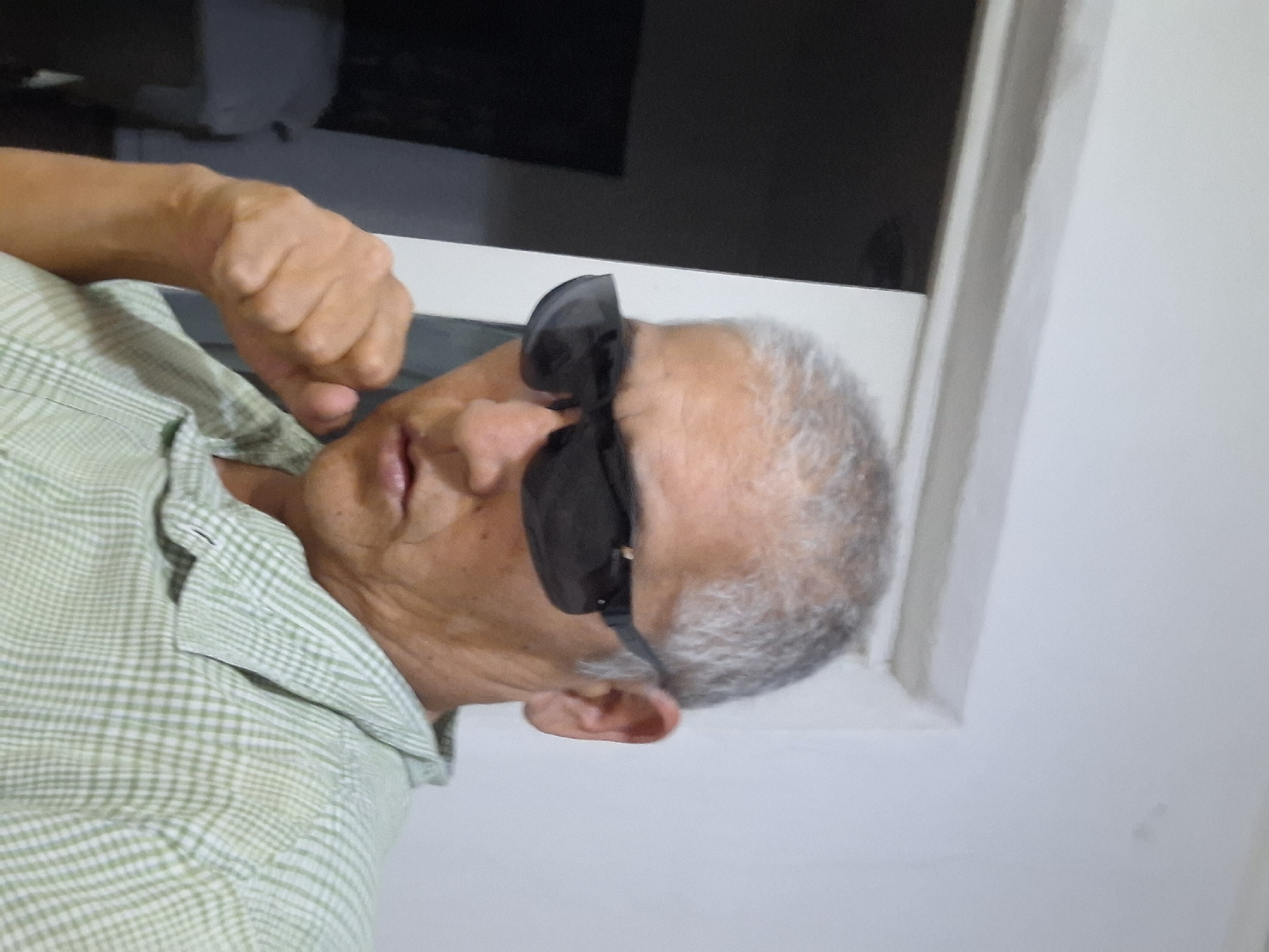 A man with short, graying hair and sunglasses is looking at the camera. He is wearing a light green and white checkered shirt. His left fist is raised near his face. He is in an indoor setting with a white wall and a doorway visible behind him. The location is not specified as Almaty, Kazakhstan.
