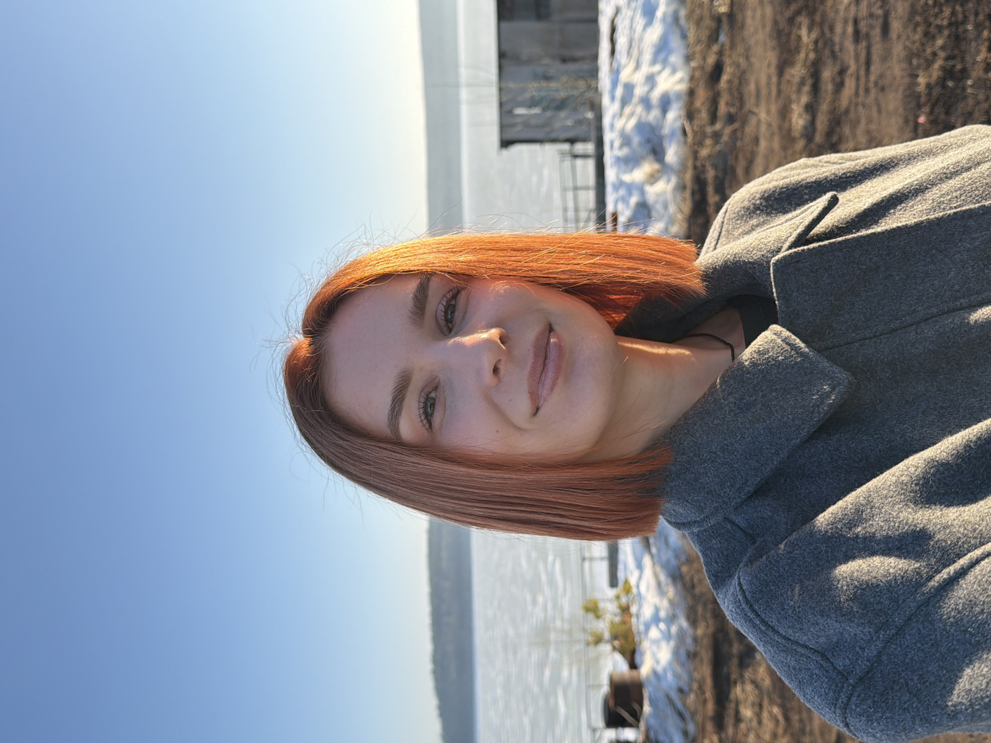 The image is a selfie of a young woman with reddish-brown hair, wearing a grey wool coat. She is smiling slightly. The background shows a body of water, likely a river or lake, and a shoreline with snow. The location appears to be near a pier or dock. Given the prompt's context, it is likely that the location is in or near Chaikovsky, Russia.