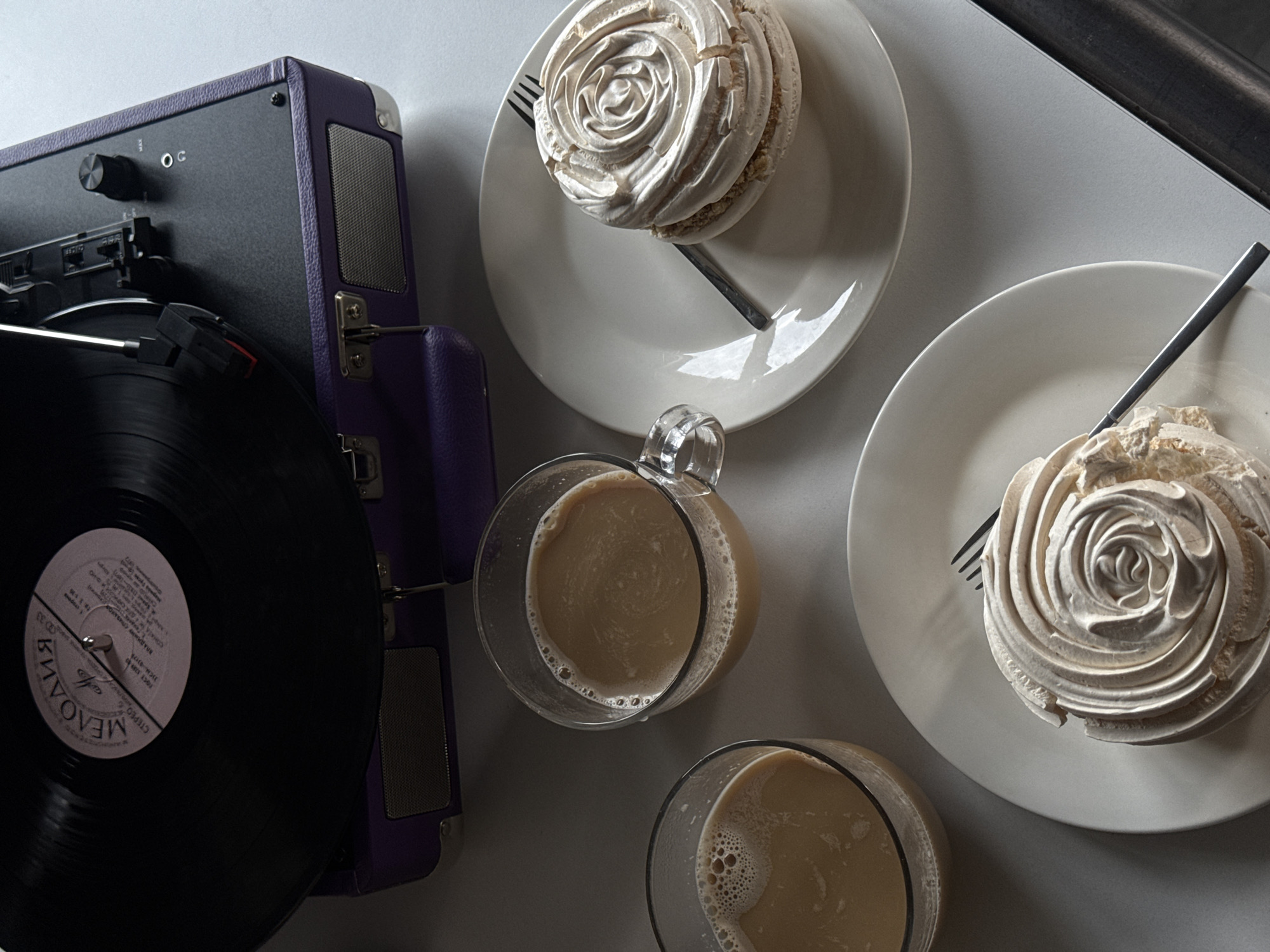 A top-down, high-angle shot presents a scene with a vintage-style turntable and two plates of dessert alongside two glasses of coffee. The turntable, primarily black with purple trim, is located on the left side of the frame. A black vinyl record rests on it. Two plates, each with a meringue-based dessert resembling a rose, are arranged towards the upper right. A fork accompanies each plate. Between the turntable and the desserts are two glasses filled with coffee, one featuring a handle. The scene is set on a plain, likely light-gray or white, surface. The image lacks discernible indicators to suggest the location of Yerevan, Armenia.