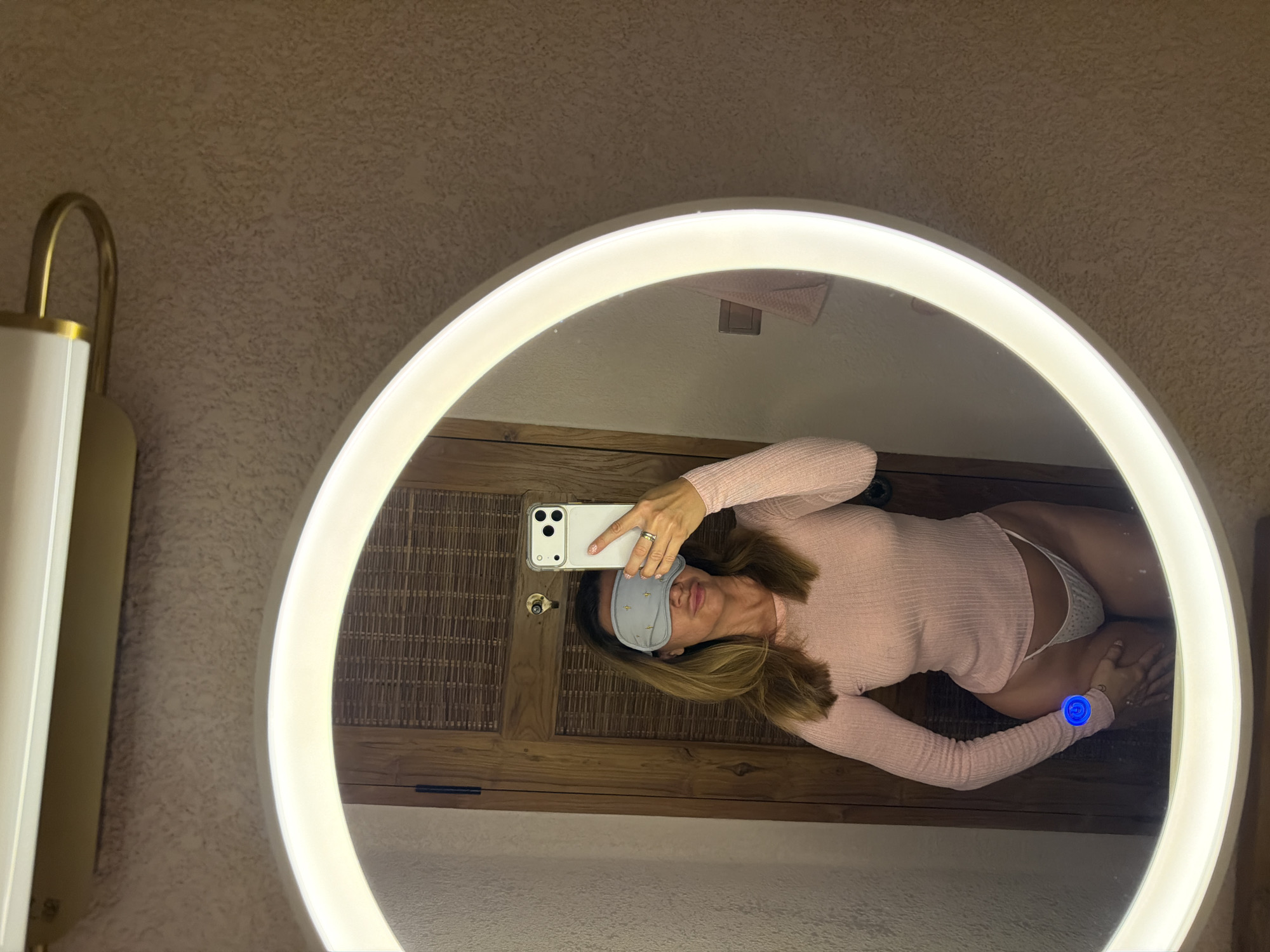 A mirror selfie taken by a woman is reflected in a circular, illuminated mirror. The woman wears a long-sleeved pink top, a sleep mask on her forehead, and a white bottom. She holds a phone with her left hand to take the picture. The background features a wooden-textured wall with a cabinet. The environment appears to be a bathroom with a light fixture visible on the left. The image appears to be taken inside a hotel room in Mengwi, Indonesia, because there is a blue light on her arm, indicating a GPS location pin was placed on the image.