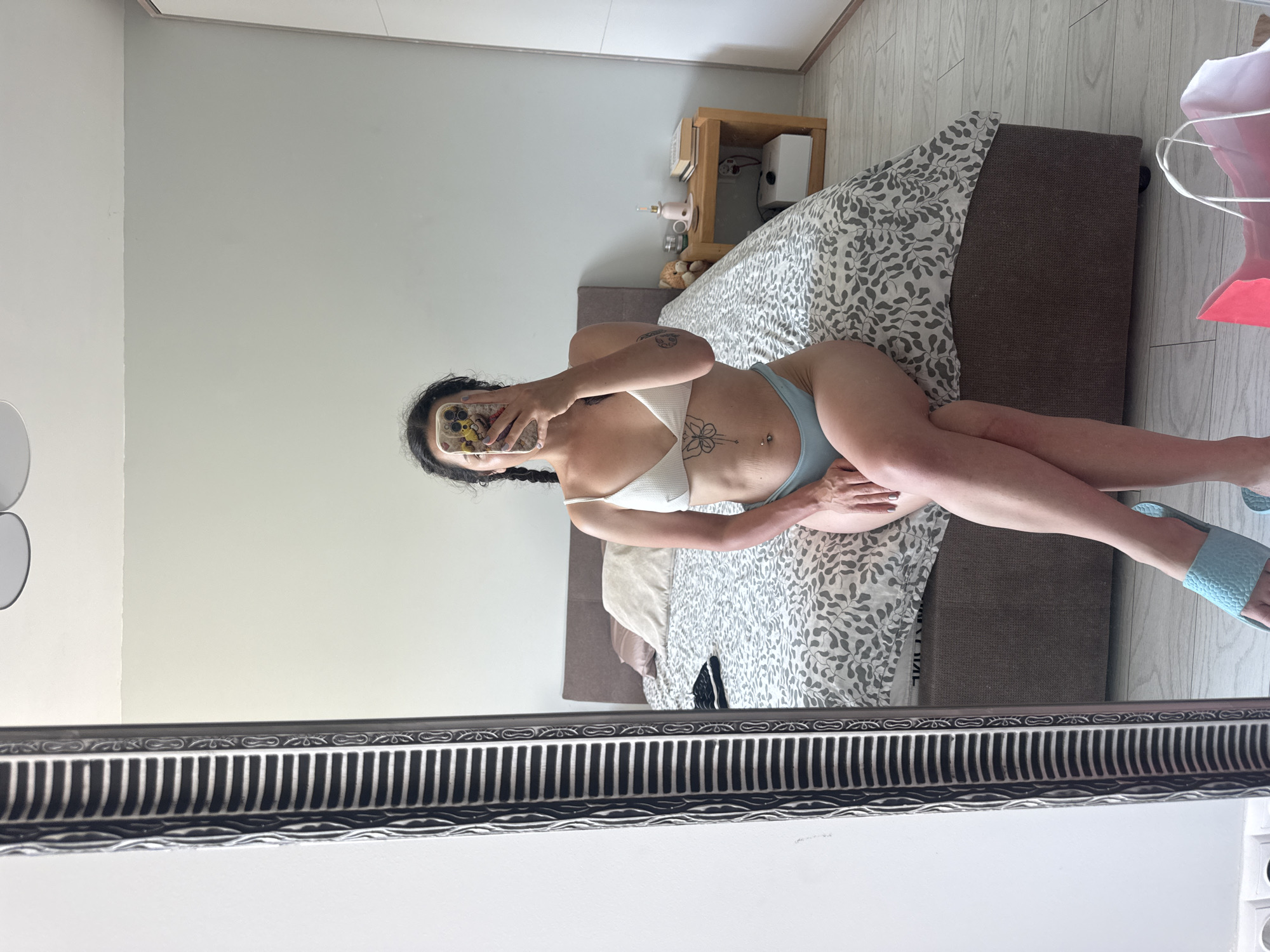 A person is taking a mirror selfie in a bedroom. They are lying on a bed with a patterned duvet, wearing a white bikini top and light blue bikini bottoms. The person has dark hair styled in braids and has tattoos on their arm and stomach. They are holding a smartphone, reflecting in the mirror. The room has light-colored walls and wooden flooring. A bedside table with decorative items and a lamp is visible. A framed mirror with an ornate design is in the foreground, obscuring the bottom portion of the scene. The lighting suggests daytime. The location is Burgas, Bulgaria.