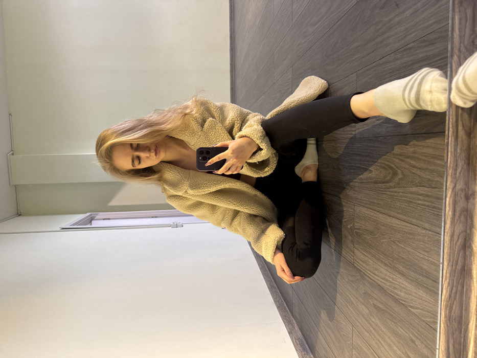 A young adult female is depicted in a mirror selfie, seated on a dark wood-grain laminate floor within an indoor room. She wears a cream-colored faux fur jacket, a dark top visible beneath, black leggings, and light-colored ankle socks with grey stripes. Her left hand holds a dark smartphone, pointed towards the viewer, while her eyes are partially closed. The indoor environment features a light beige wall on the left with a white vertical conduit pipe, and a light green wall on the right. A partially visible dark doorway with a light frame is on the left. The exact location within the city of Unknown, Unknown is not determinable from the image; the setting appears to be a functional interior space, possibly a studio or commercial hallway.