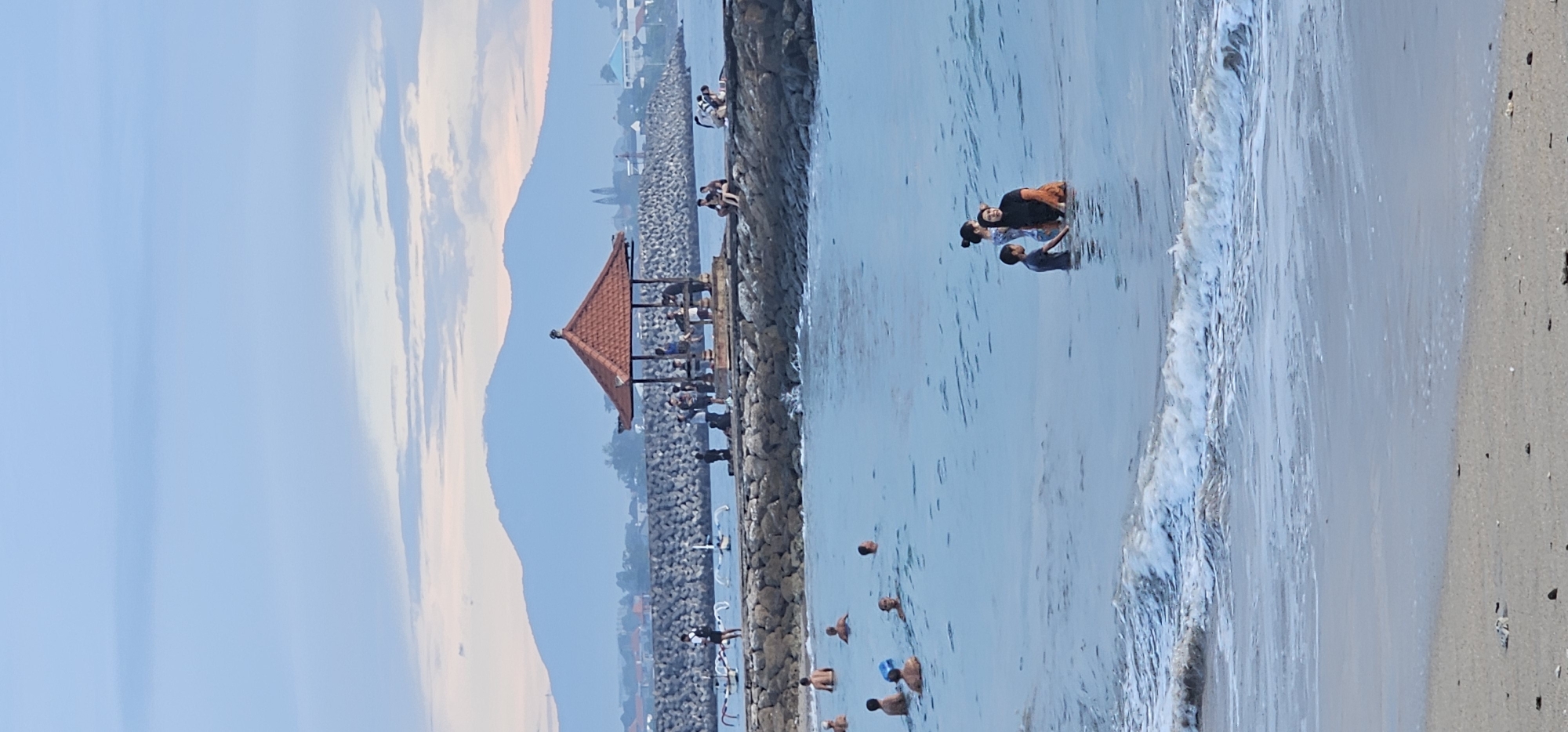 This photograph captures a serene yet active coastal scene in Denpasar, Indonesia, likely during sunrise or sunset, indicated by the soft, warm light and pastel hues in the sky.
The image is horizontally composed, with a significant portion dedicated to the ocean and a long, rocky pier or breakwater extending into the water. In the mid-ground, a prominent traditional-style gazebo with a tiered, red-orange tiled roof stands on this pier. Several people are gathered under or around the gazebo, and more individuals are seen along the length of the stone pier, sitting and observing. Beyond the pier, distant buildings line the coastline.
The background is dominated by large mountains or hills, silhouetted against a pale blue sky streaked with soft white and pinkish clouds.
In the foreground and to the right, the ocean stretches out, its clear, light blue waters showing gentle white-capped waves approaching a sandy beach. Numerous people are enjoying the water, wading and swimming. A group of three individuals – two adults and a child – are prominently visible standing in the shallow water. Other individuals are further out in the sea. The sandy beach on the far right is light-colored and appears somewhat coarse, with small stones scattered.
The overall scene depicts a leisurely atmosphere, with people enjoying the natural beauty and recreational opportunities of the Indonesian coast. No visible text is present in the image.