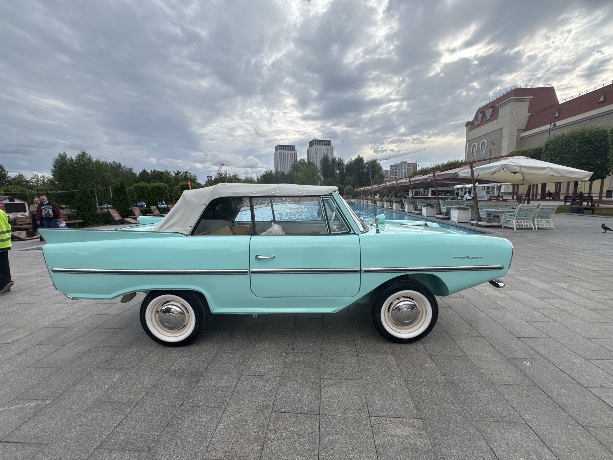 A vintage light blue convertible Amphicar is parked on a paved area with a pool in the background. The car has a white convertible top and white-walled tires. Two people are visible in the distance to the left, near a small building and some trees. Behind the car, there is a swimming pool, followed by a row of tables and chairs under umbrellas. A large building with a red roof and arched windows is situated further in the background, along with several modern high-rise buildings and a construction crane. The sky is overcast with scattered clouds. The location is identified as the Amphibious Car Museum in Moscow, Russia.