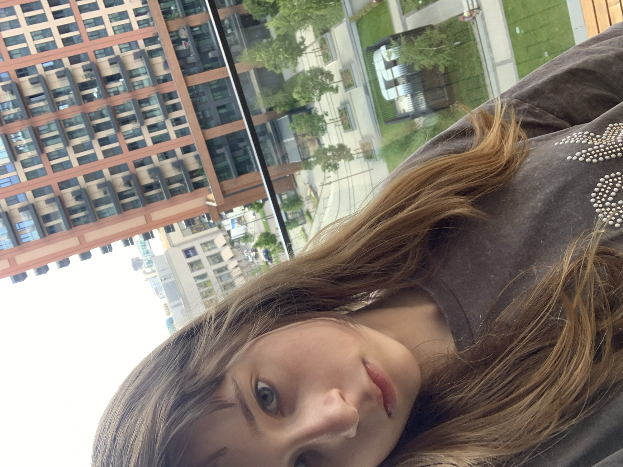 A selfie-style photograph features a light-skinned individual with long, light brown hair. The person is wearing a dark gray t-shirt with a studded star design on the shoulder. The background reveals an urban landscape visible through a window, featuring tall brick and glass buildings, a park with trees, and streetscape elements. The location is Belgrade, Serbia.