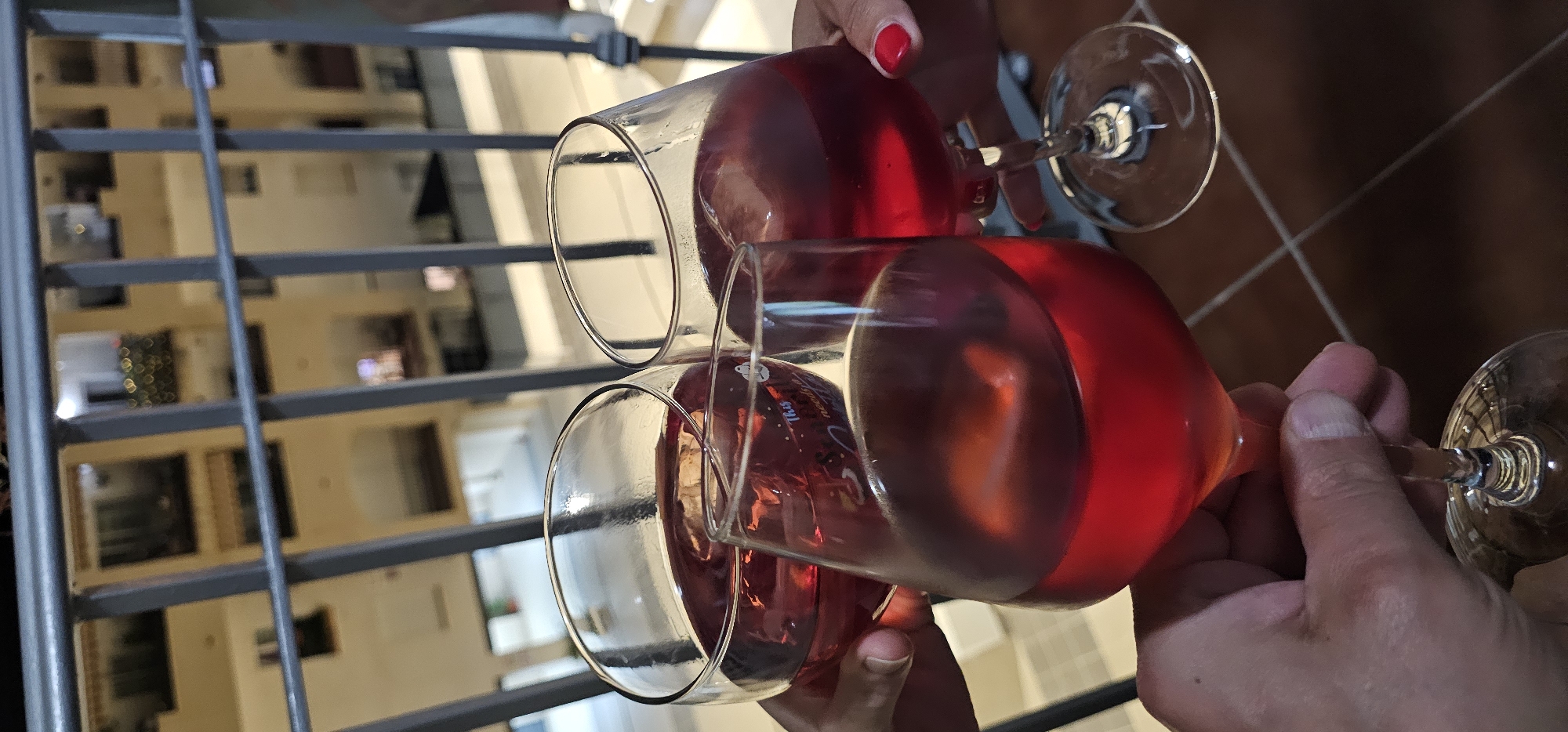 Two individuals are captured in the act of toasting with wine glasses, seen from a first-person perspective, possibly on a balcony or terrace in Manilva, Spain. Two hands are visible, one adorned with red nail polish holding a glass on the right, and another hand with shorter nails holding a glass on the left, as the glasses clink together. Both stemware glasses are filled with a translucent, reddish liquid, likely red wine or sangria.
The background, visible through a dark grey metal railing, features a blurred view of a multi-story building, suggesting an apartment complex or hotel. Some lights are visible in the background building, creating a bokeh effect, indicating the photo was taken at dusk or night. A tiled floor, dark brown with lighter grout lines, is partially visible in the lower right portion of the frame.
On the glass held by the hand with shorter nails, faint white text is visible, including the word "This" and "mejores" (Spanish for "best"), with "16:35" printed beneath it. The scene conveys a relaxed and celebratory atmosphere.