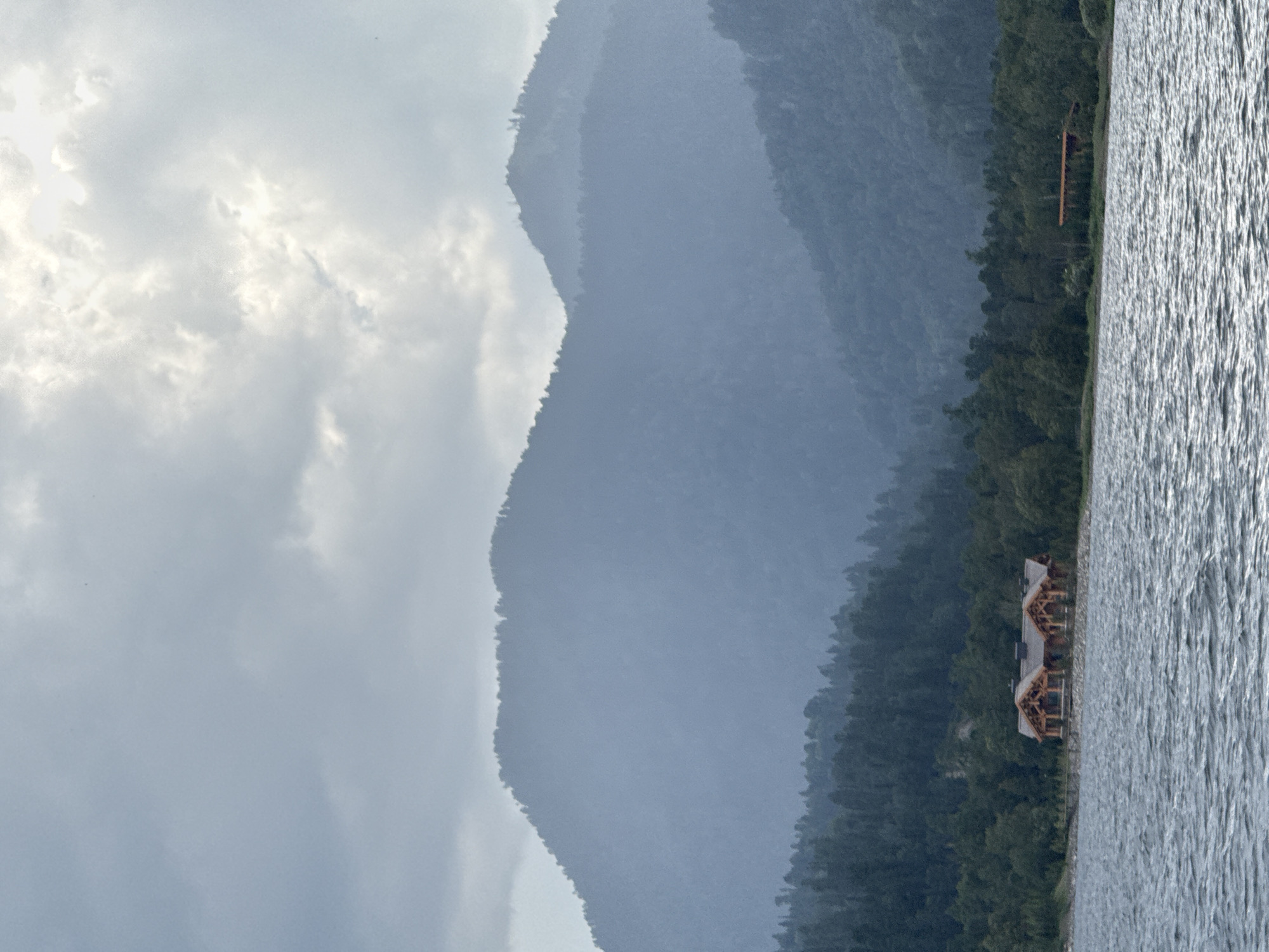 A lakefront scene at Kordon Baigazan, Russia, under an overcast sky. In the foreground, choppy, dark gray water reflects the muted light. Along the shoreline, a rustic, wooden lodge-style building with multiple gables and exposed beams stands prominently. Behind the lodge, a dense forest of dark green trees covers the lower slopes of misty, blue-gray mountains that dominate the background. The peaks of the mountains are softened by atmospheric haze. To the right of the lodge, a smaller, reddish-brown structure is partially visible amongst the trees. The overall impression is one of serene, natural beauty, with a touch of rugged human habitation.