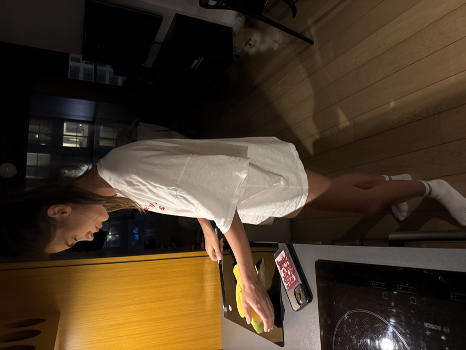 A woman in a white t-shirt and socks stands in a kitchen, slicing a yellow object on a black cutting board. A cell phone with a decorative case sits nearby. The kitchen features a wooden counter, stove, and cabinets. A window reflects city lights. A dog is visible in the background. The floor is wooden.