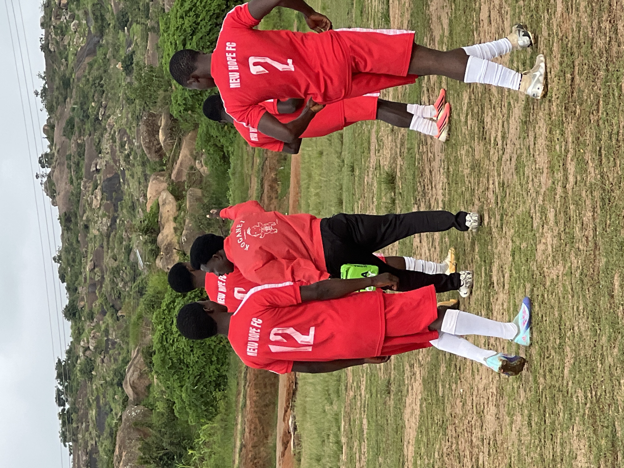 In Jos, Nigeria, a group of young men, likely a football team, are gathered on a dusty, grass-covered field. The sky is overcast, and in the background, a rugged, rocky hillside is covered in green vegetation. The men are wearing red jerseys and shorts with white trim. One player in the foreground has the number "12" on his back, while another has the number "2". The jerseys have "NEW HOPE FC" written on them. One man, possibly a coach, is wearing a red jacket with a graphic and "KOGANEl" written on it. The players appear to be standing and interacting casually. The field itself shows signs of wear and tear, with patches of bare earth visible.