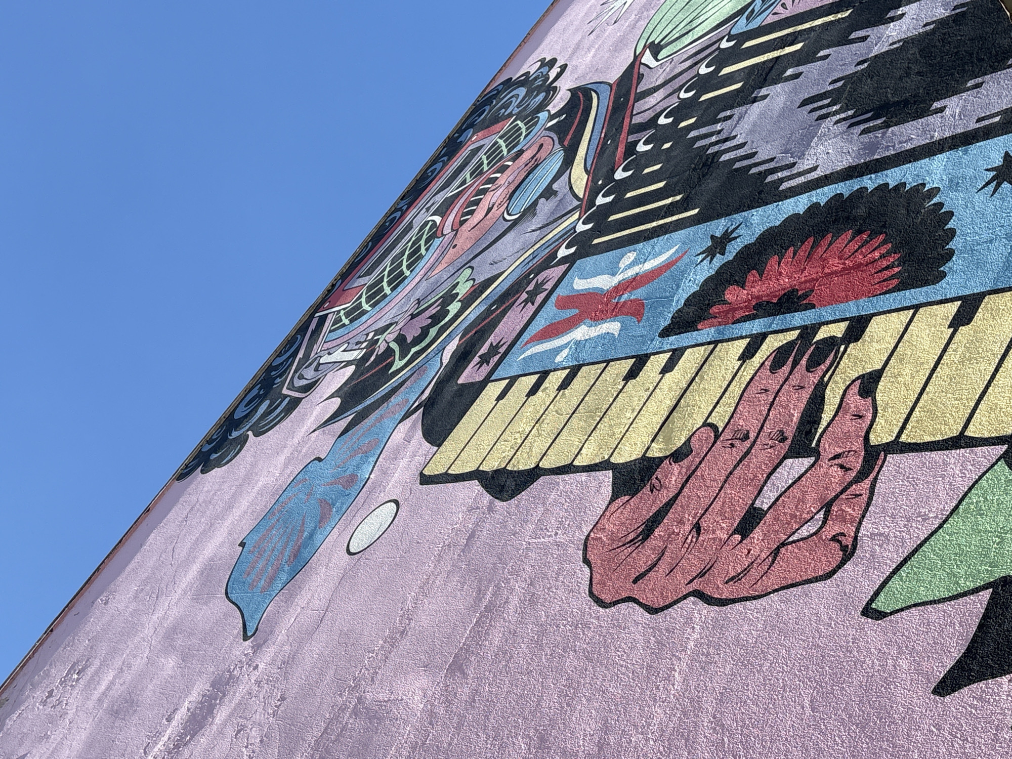 The image shows a close-up of a colorful mural painted on a textured pink wall, seen from a low angle against a clear blue sky. The mural features abstract, psychedelic imagery with bold lines and vibrant colors. Prominently displayed are a large, stylized red hand with fingers extended, positioned over a section of yellow and black piano keys. To the right of the hand and keys, other abstract shapes and figures are visible, including a bird-like figure in red and blue, and patterns of black, purple, and green. The mural appears to be on the side of a building, suggesting an urban setting. This mural is located on the side of the SESC Pompeia building in São Paulo, Brazil.