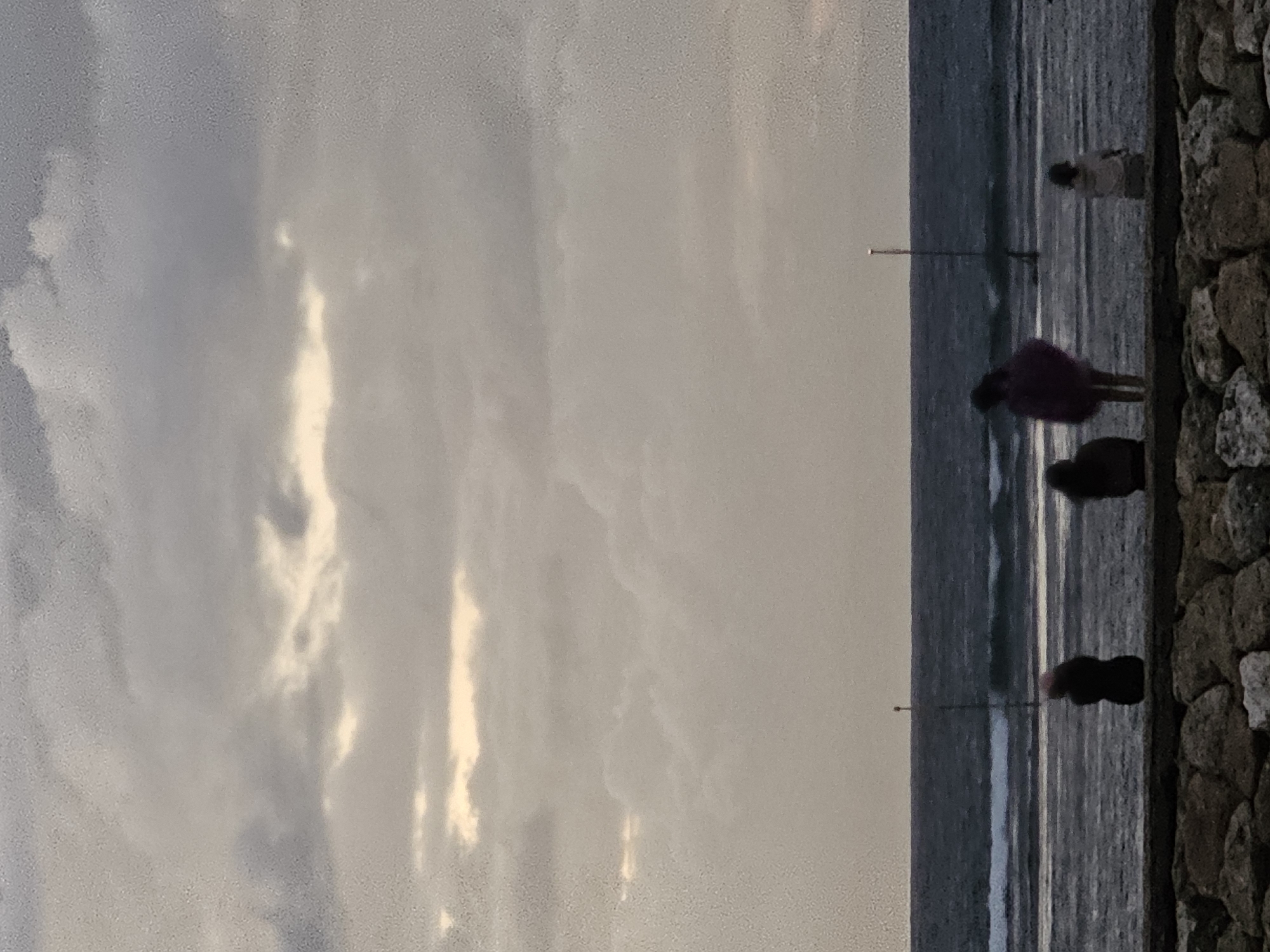 The image depicts a cloudy sky over a choppy ocean. In the foreground, a low stone wall borders a wet, sandy beach. Four individuals are visible on the beach. From left to right, the first person is silhouetted against the water. The second person wears a purple garment and appears to be standing. The third person is a smaller figure. The fourth person is also silhouetted. Two tall poles with small markers at the top are in the water, one near the horizon and one closer to the shore. The overall scene is overcast, suggesting a moody atmosphere. This location is on the coast of Denpasar, Indonesia.