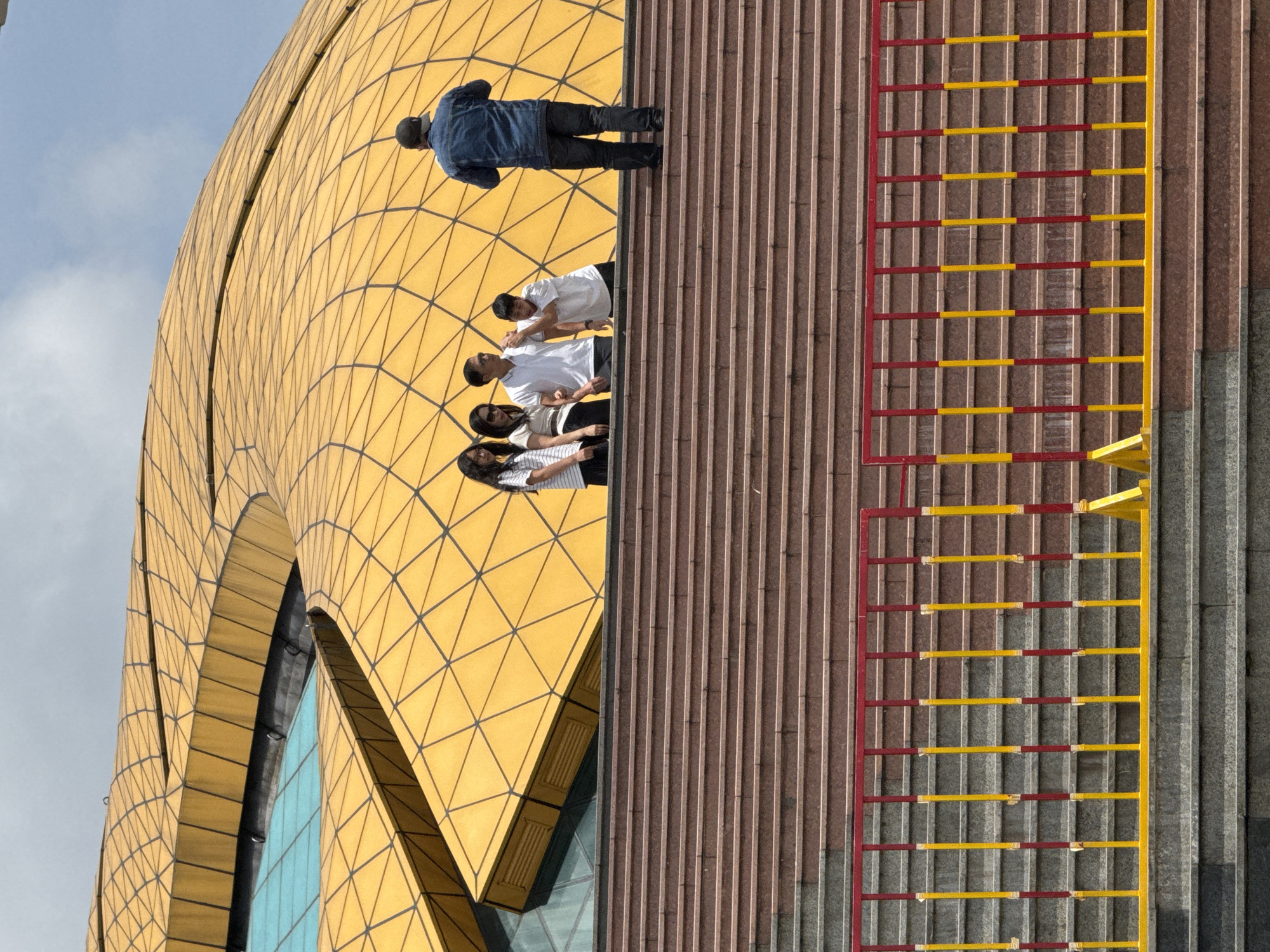 A group of five individuals, four younger people and one adult male, stand on a wide, tiered concrete staircase. The staircase is lined with red and yellow metal railings. Behind the individuals, a large, undulating yellow structure with a geometric pattern, possibly a building roof or facade, dominates the background. The sky above is blue with scattered clouds. The scene appears to be outdoors in an urban setting, likely a public space such as a park or cultural center. This location is the Da Lat Pedagogical College, also known as the Da Lat Teacher Training College, in Da Lat, Vietnam.
