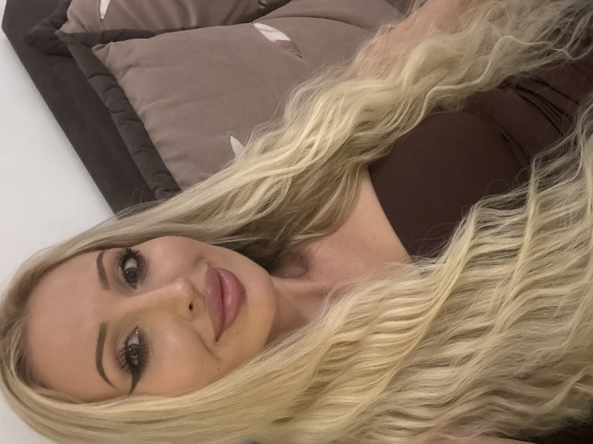 A young woman with long blonde hair, wearing a dark brown strapless top, is pictured indoors, leaning against a dark gray sofa with taupe cushions. Her makeup includes winged eyeliner and pink lipstick. The lighting suggests an indoor environment, possibly a living room or bedroom. The background consists of the sofa and a white wall.