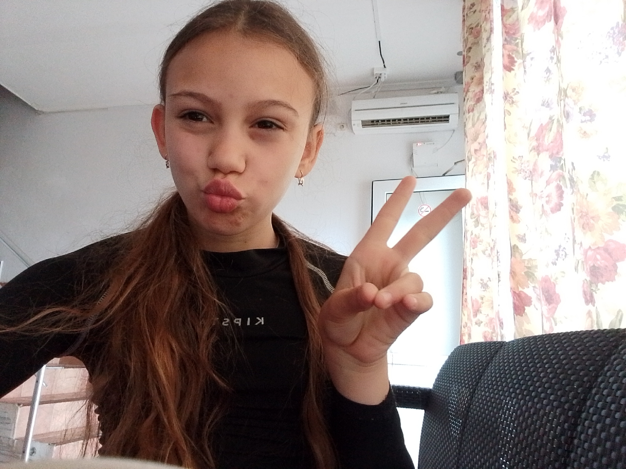 A young girl with long brown hair, parted in the middle and pulled back into two low ponytails, is taking a selfie. She is wearing a black long-sleeved shirt with white writing on it and silver hoop earrings. She is making a kissing face while holding up a peace sign with her right hand.
Behind her, there is an air conditioning unit mounted on the wall. To the right, there are floral curtains. A black mesh chair is visible in the lower right corner. A non-smoking sign is also visible.
The location is likely an indoor space in Sochi, Russia.