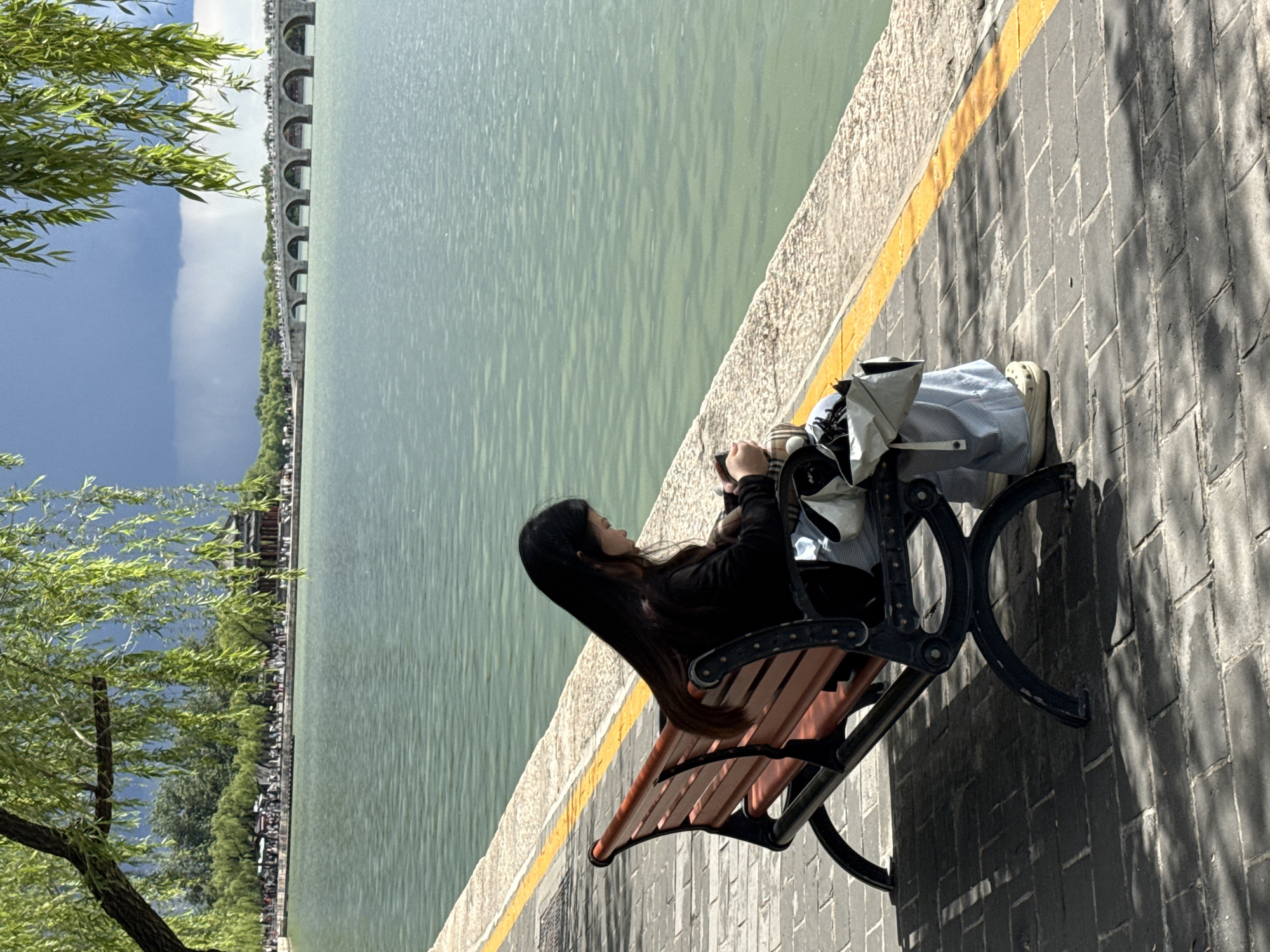 A young woman with long, dark hair is seated on a park bench beside a large body of water in the Haidian District of China. She is wearing a dark long-sleeved top and light-colored pants, and appears to be looking down at something in her hands, possibly a phone. A light-colored bag or garment is resting beside her on the bench. The bench itself has a black metal frame and wooden slats, casting shadows on the paved walkway where it sits.
The paved walkway, made of rectangular bricks, has a prominent yellow line running along its edge, separating it from a stone barrier next to the water. The water, likely a lake or large pond, is a muted greenish-blue and shows ripples on its surface. In the background, across the water, a long arched bridge is visible, with numerous arches spanning the width. Beyond the bridge, there's a line of lush green trees, and further in the distance, a blue sky with some cloud formation.
On the left side of the image, the leaves of a tree, possibly a willow, are prominently displayed, indicating a sunny day with dappled light creating shadows on the walkway. The overall atmosphere suggests a peaceful outdoor setting. No visible text is present in the image.