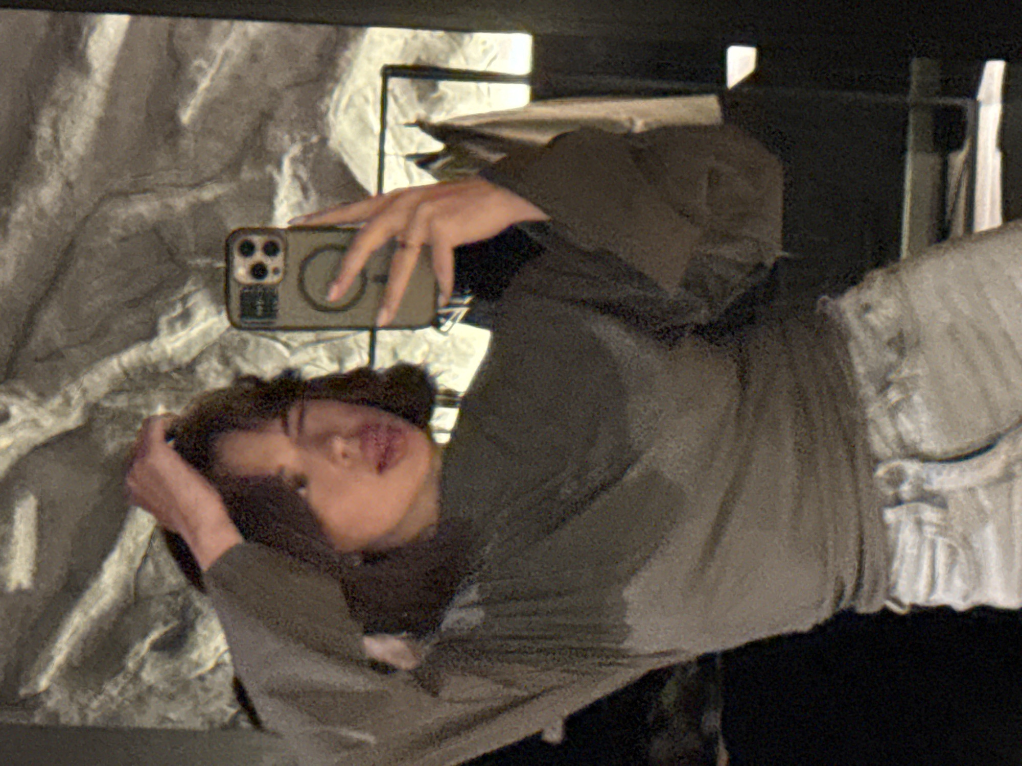 A woman is taking a selfie in front of what appears to be a decorative wall panel. She's holding a phone with a distinctive camera array and a case featuring a circular design. The woman is wearing a dark-colored top with wide sleeves and light-colored bottoms. Her hair is brown. The setting includes elements of interior design, like black framing and reflections in what looks like a mirrored surface. There is no evidence to connect the image with the city of Chaikovsky, Russia.