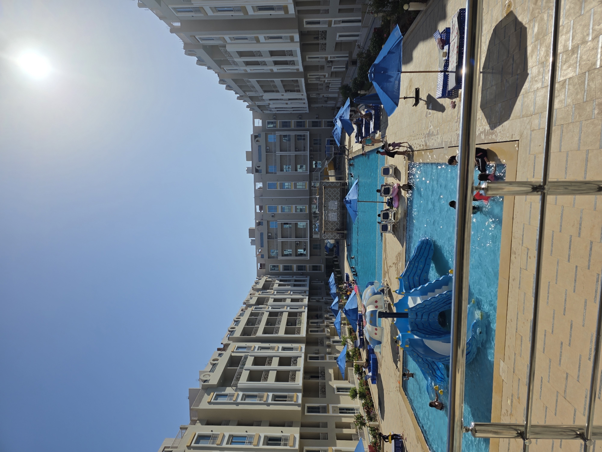 The image shows a swimming pool area in Al Hadaba, Egypt. Two multi-story buildings flank the pool on opposite sides. Blue umbrellas and lounge chairs are situated along the pool deck. People are swimming and interacting in the pool. A blue water slide is positioned at one end of the pool. The sky is clear with the sun visible. A metal railing is in the foreground.