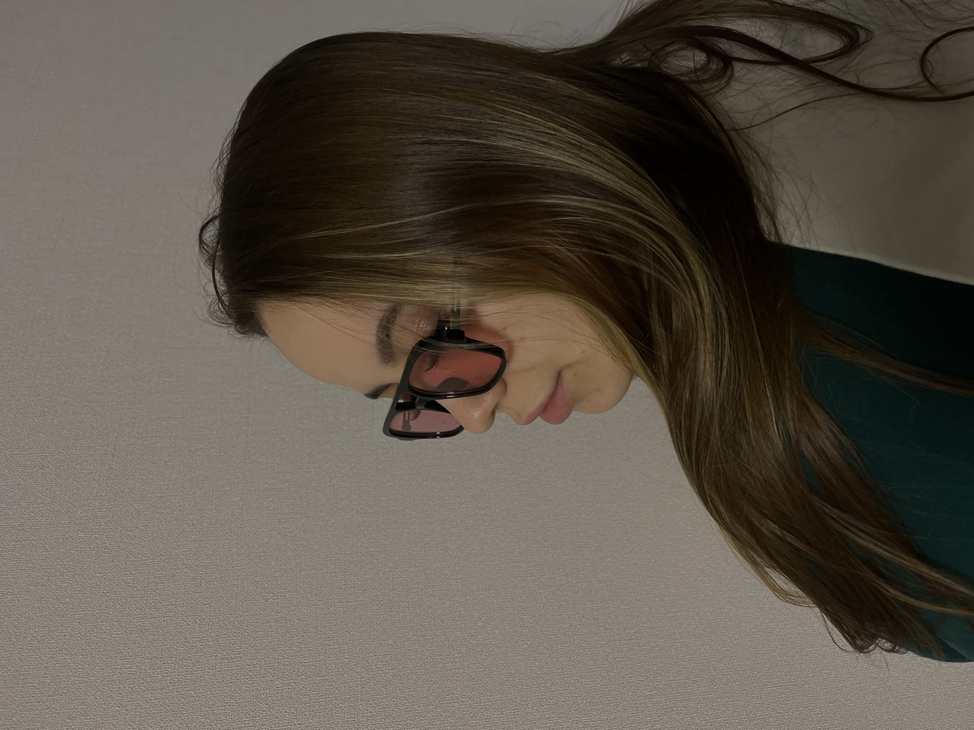 A photograph shows a woman with long, brown hair wearing pink-tinted sunglasses. She is wearing a dark green garment. The background is a plain, light grey wall. The location is not discernible from the image, and there is no indication the image was taken in Krasnodar, Russia.