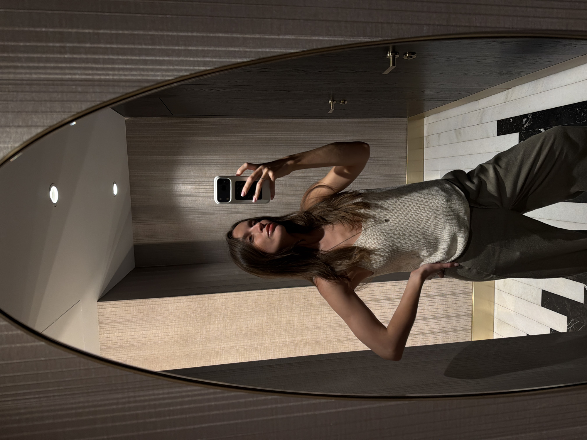 A woman with long brown hair holds a cell phone and poses in a mirror. The mirror reflects her image and the surrounding bathroom, which features a wall with horizontal striped paneling, a countertop with a white and black tile backsplash, and brass fixtures. The location is a bathroom in Budva, Montenegro.