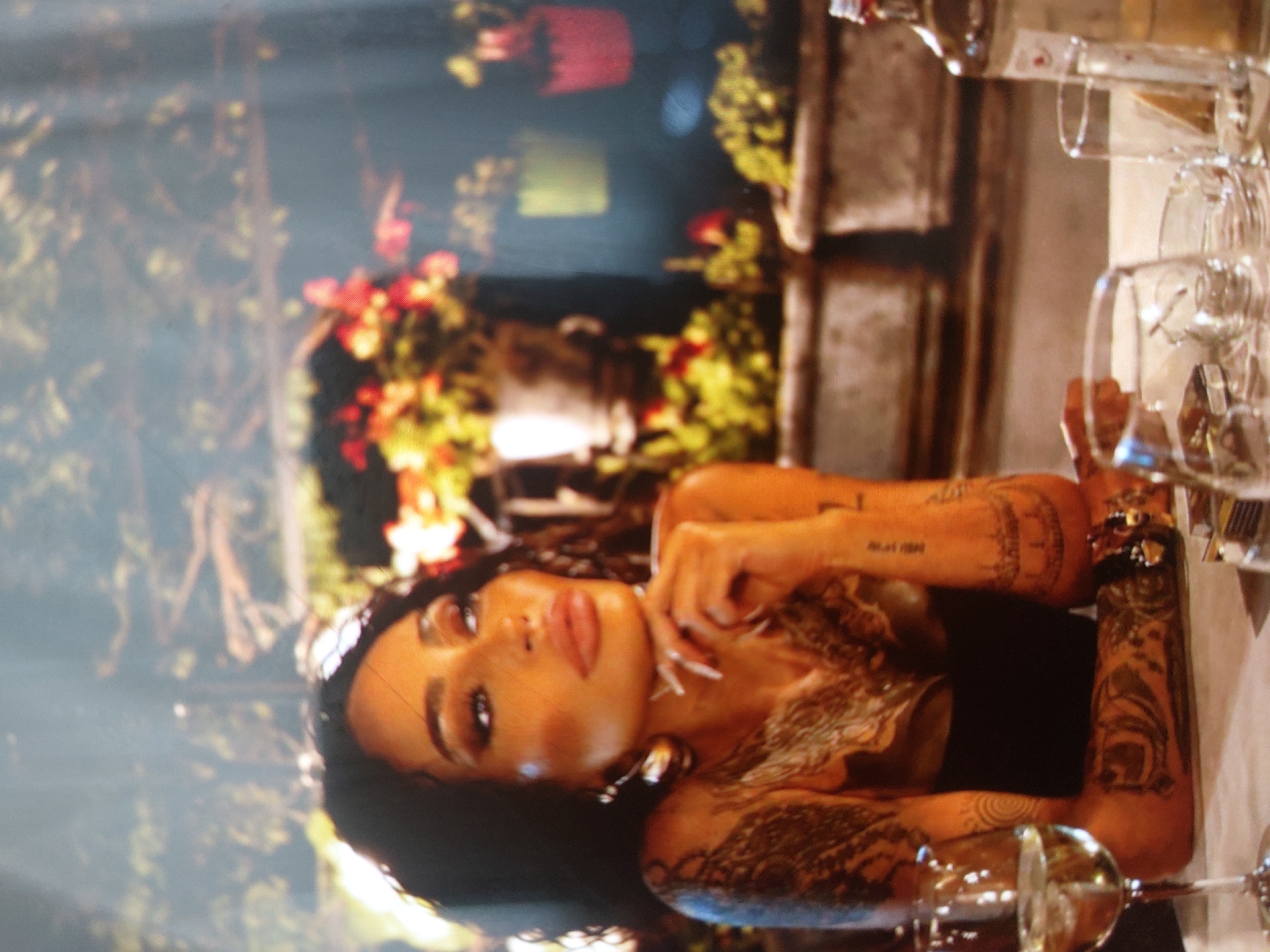 A medium shot depicts a woman with dark hair and multiple tattoos on her arms lying in a tub or body of water surrounded by flowers and greenery. The environment seems to be outdoors, possibly a garden or patio, with architectural elements like stone planters visible in the background. Glasses, some seemingly stacked, are also visible, suggesting a table setting. The woman is the central focus, positioned near the bottom of the frame. Given the floral arrangement and overall composition, this appears to be a staged or carefully arranged scene. There is no information in the image to suggest that it was taken in Tsarevo, Bulgaria.