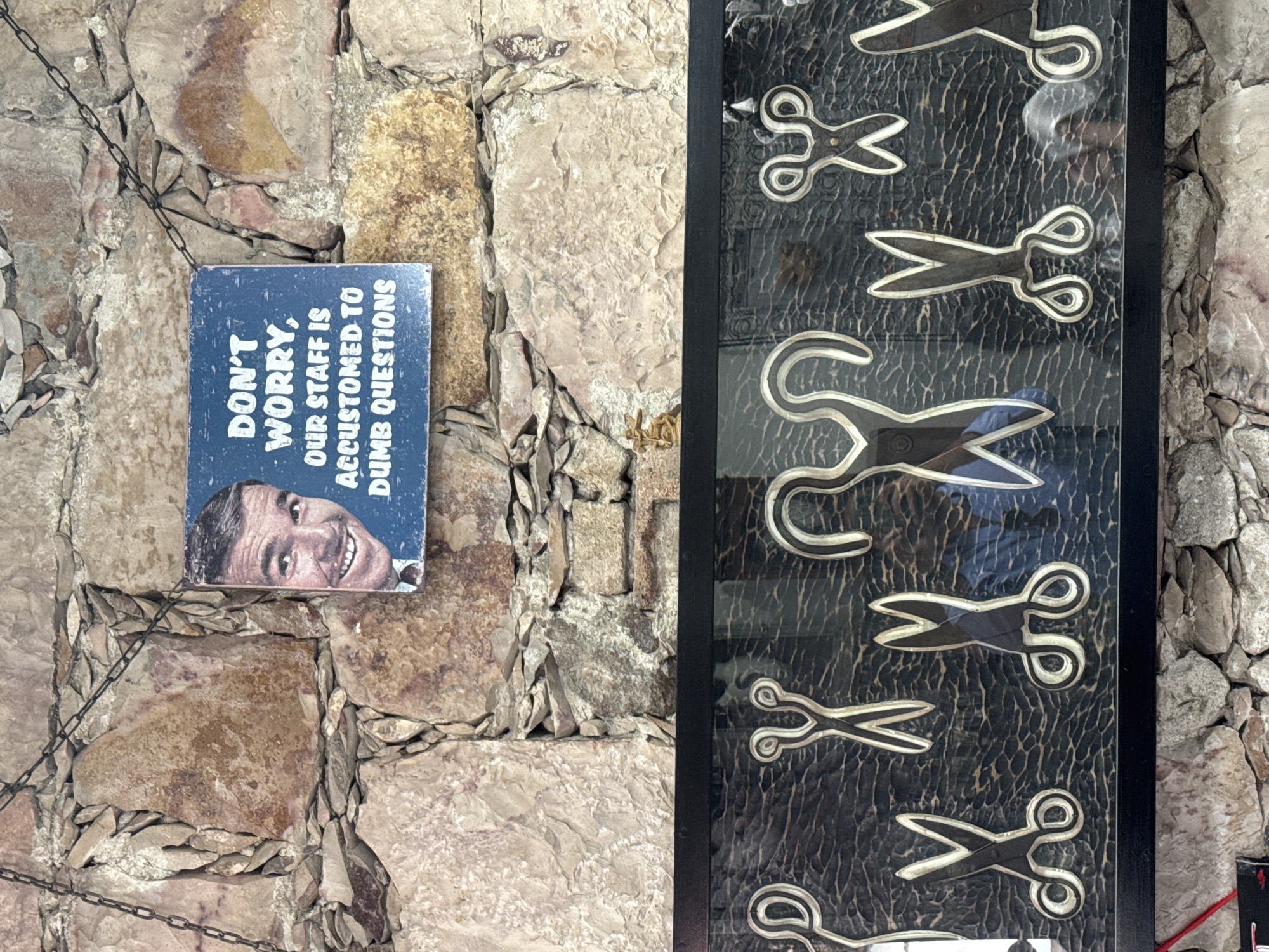 The image shows a stone wall on which a sign reading "DON'T WORRY, OUR STAFF IS ACCUSTOMED TO DUMB QUESTIONS" is displayed along with a picture of a smiling man, next to a framed display of decorative scissors. This likely exists in the city of San Miguel de Allende, Mexico, suggested by architectural features.