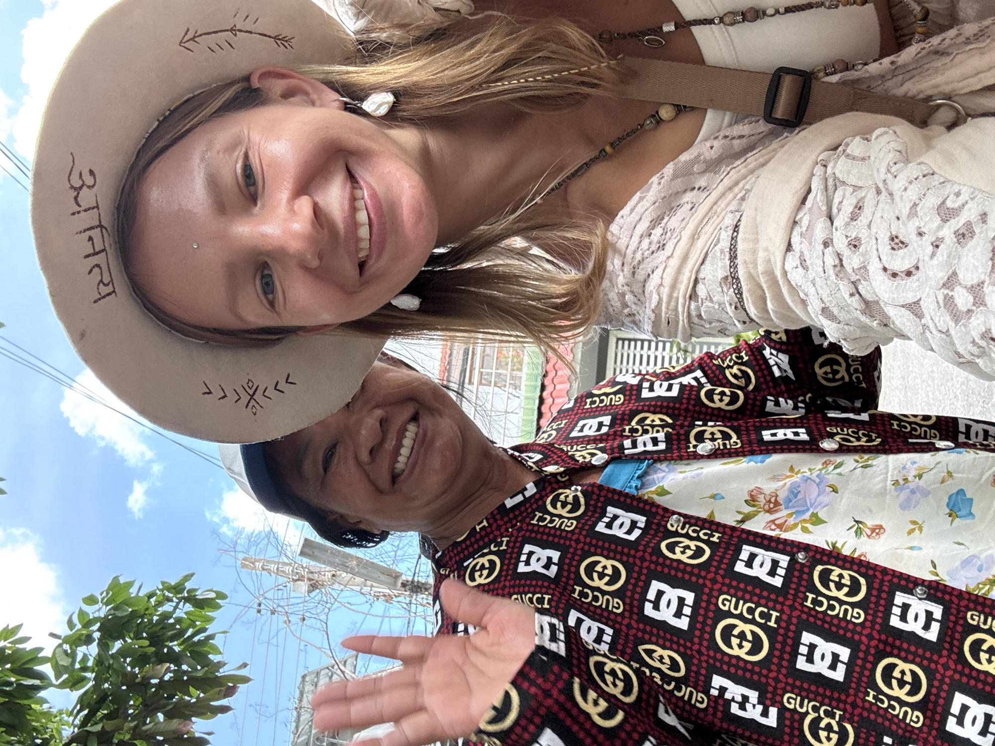 In Phu Thuy Ward, Vietnam, two women are taking a selfie outdoors on a sunny day. The woman on the left, wearing a white and blue cap and a patterned red and black jacket with "GUCCI" branding, is smiling and waving her left hand. She is also wearing a floral print blouse. The woman on the right, with blonde hair, is wearing a wide-brimmed hat with decorative markings, earrings, and a lace shawl draped over a white top. She has a septum piercing and is also smiling. The background shows a cloudy blue sky, a tree with green leaves, and some electrical wires and poles.