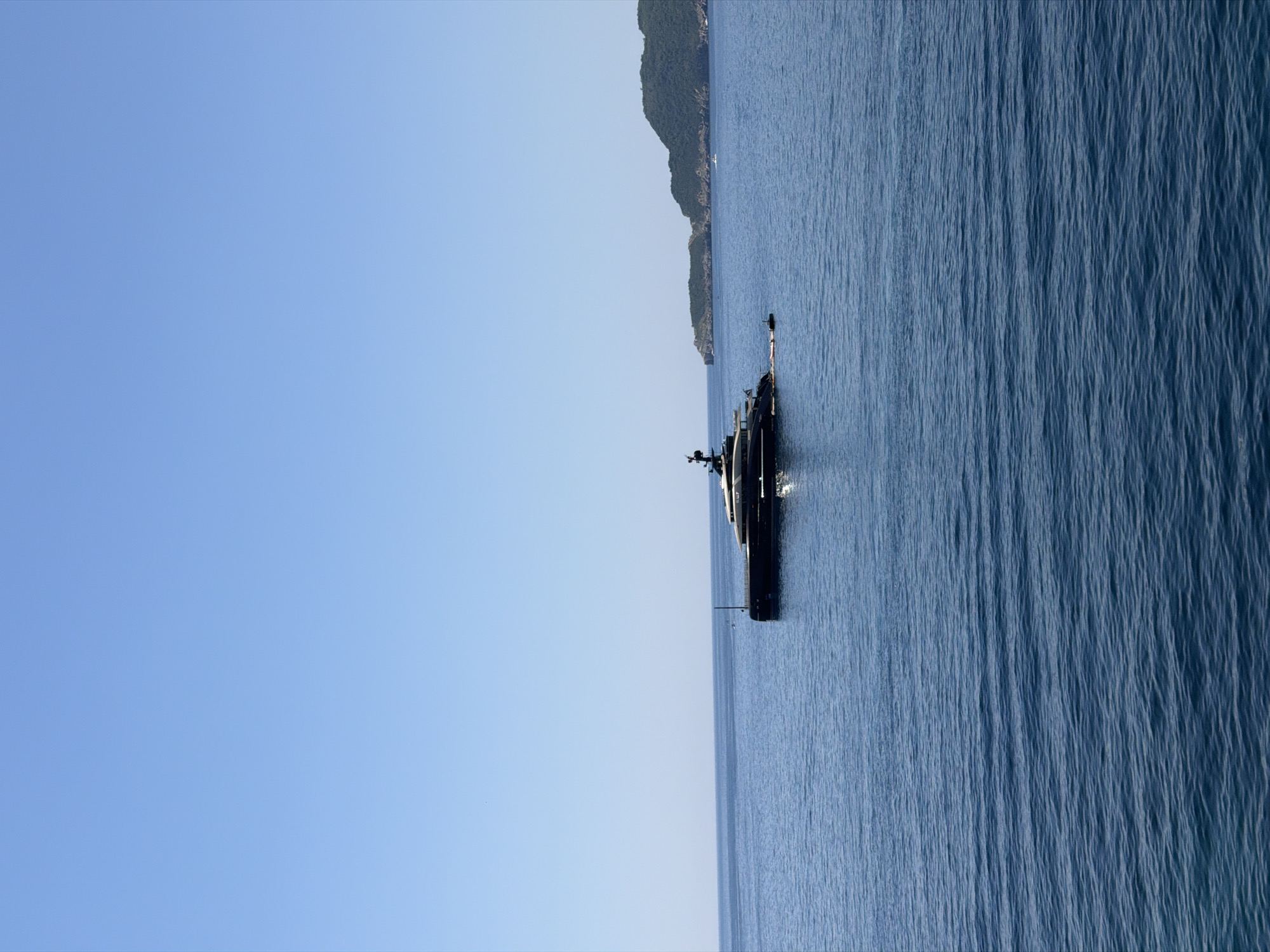 An aerial, wide-angle shot captures a sleek, black superyacht anchored in the azure waters off the coast of Rađenovići, Montenegro. The vessel, characterized by its modern design and multiple decks, sits prominently in the frame, surrounded by calm, rippling sea. In the distance, a verdant, mountainous island coastline lines the horizon, its lush greenery contrasting with the clear, cloudless blue sky. The scene evokes a sense of luxury and tranquility.