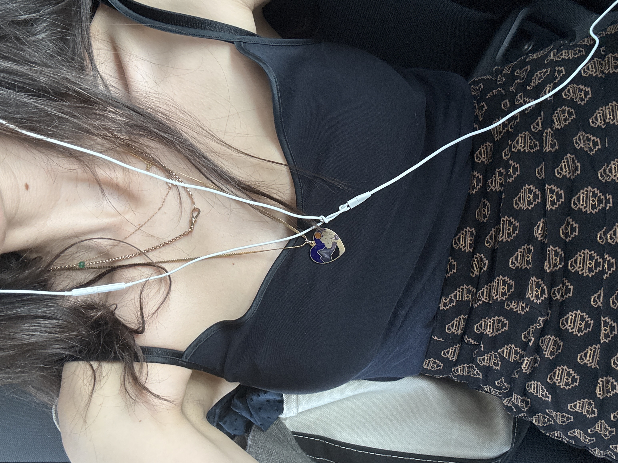 This is a close-up, overhead shot of a person from the chest down. The person is wearing a black tank top and a patterned skirt with a dark background and a repeating beige geometric design. They have long, dark brown hair that falls over their shoulder. The person is wearing multiple necklaces, including one with a heart-shaped pendant depicting a landscape scene with a sun. White earbuds are visible, with the cord draped over their chest. The background appears to be the interior of a vehicle, with a portion of a bag visible on the left. The scene is well-lit, likely with natural light. No specific New York City landmarks or indicators are present in the image.