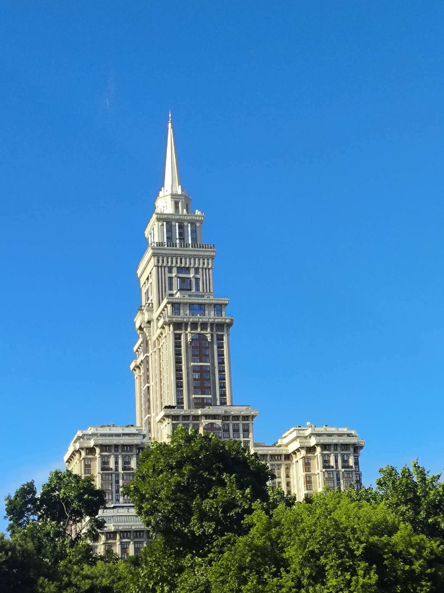 The media file is a low-angle, full shot of a grand, multi-story building with a spire, set against a clear, bright blue sky. The building is light-colored, likely stone or stucco, with numerous windows and decorative architectural details. It appears to be a prominent landmark, possibly a hotel or government building. The spire at the top is tall and slender, tapering to a point.
In the foreground, lush green trees obscure the lower portion of the building, suggesting the photo was taken from ground level within a park or heavily treed area. The trees are dense and vibrant, indicating a warm, likely summer day. The bright sky and clear visibility suggest fair weather, and the sunlight on the building indicates it's daytime, possibly late morning or afternoon. No people are visible in the media file. There is no discernible text in the image. The overall impression is one of grandeur and architectural splendor, framed by the natural beauty of the trees and the vastness of the sky.