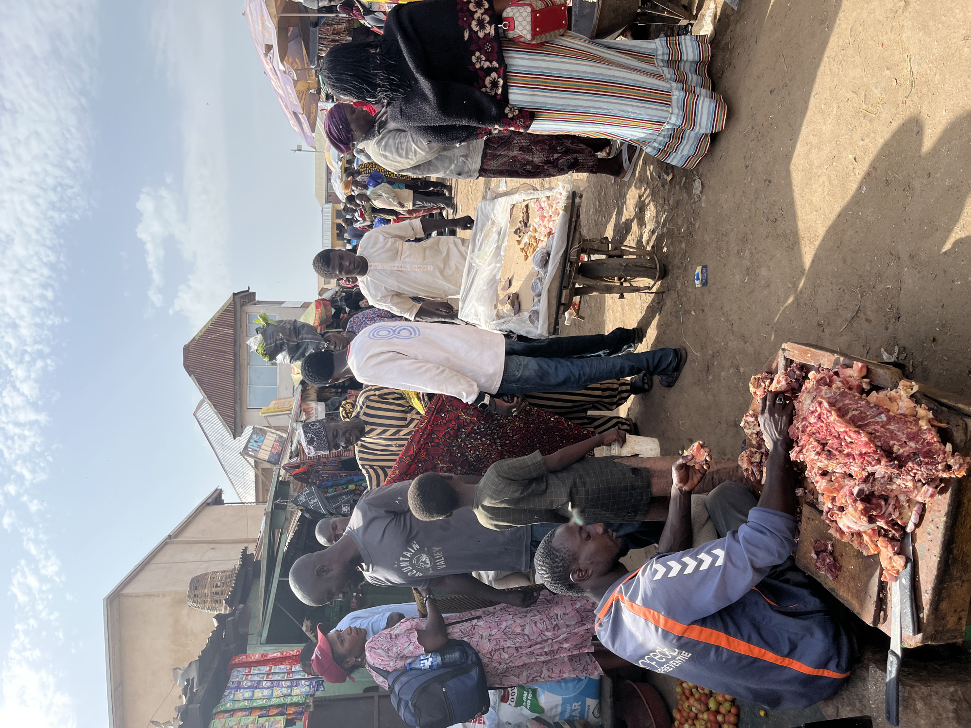 The image depicts a bustling outdoor market scene in Rafin Par, Nigeria. In the foreground, a man is seated and actively butchering a large quantity of raw meat on a wooden block, with a knife nearby. To his left, a pile of tomatoes is visible. The market is populated with numerous individuals, some engaged in transactions, others observing. To the right of the butcher, a man in a white garment displays various items on a cart covered with plastic sheeting. In the background, market stalls with corrugated metal roofs are visible, some displaying goods. The ground is unpaved dirt. The sky is partly cloudy.