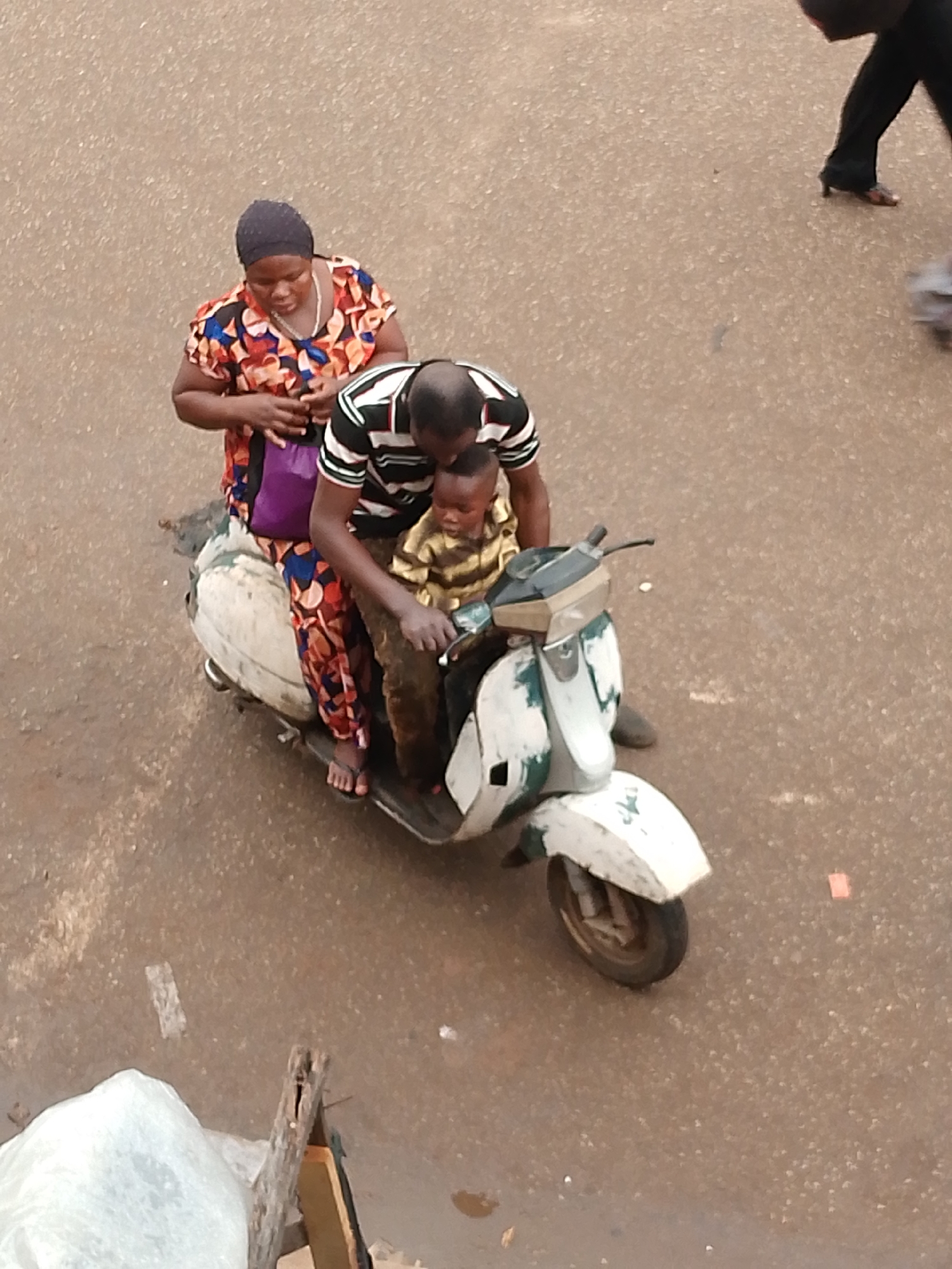 A medium, high-angle shot depicts three people on a white moped on a mottled brown road surface. The location is Bukuru, Nigeria. A dark-skinned woman with a black head covering and a colorful floral-patterned dress sits behind a dark-skinned man with a shaved head, wearing a striped black and white shirt. He is holding the handlebars. A child in a light-colored striped shirt stands in front of the man. The woman has a purple bag. An adult walks in the upper right. There is debris in the lower left.