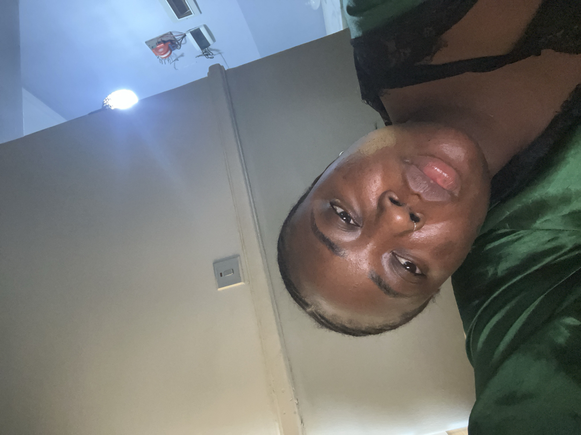 A close-up, slightly skewed, indoor selfie of a dark-skinned individual with short, braided hair. The person has a nose piercing and is wearing a dark top with lace detailing and a green, velvet-like garment. The background is a plain, light-colored wall with a light switch. The overhead ceiling has a recessed light fixture and electrical wiring exposed. There is no discernible information to establish the location as Kubwa, Nigeria.