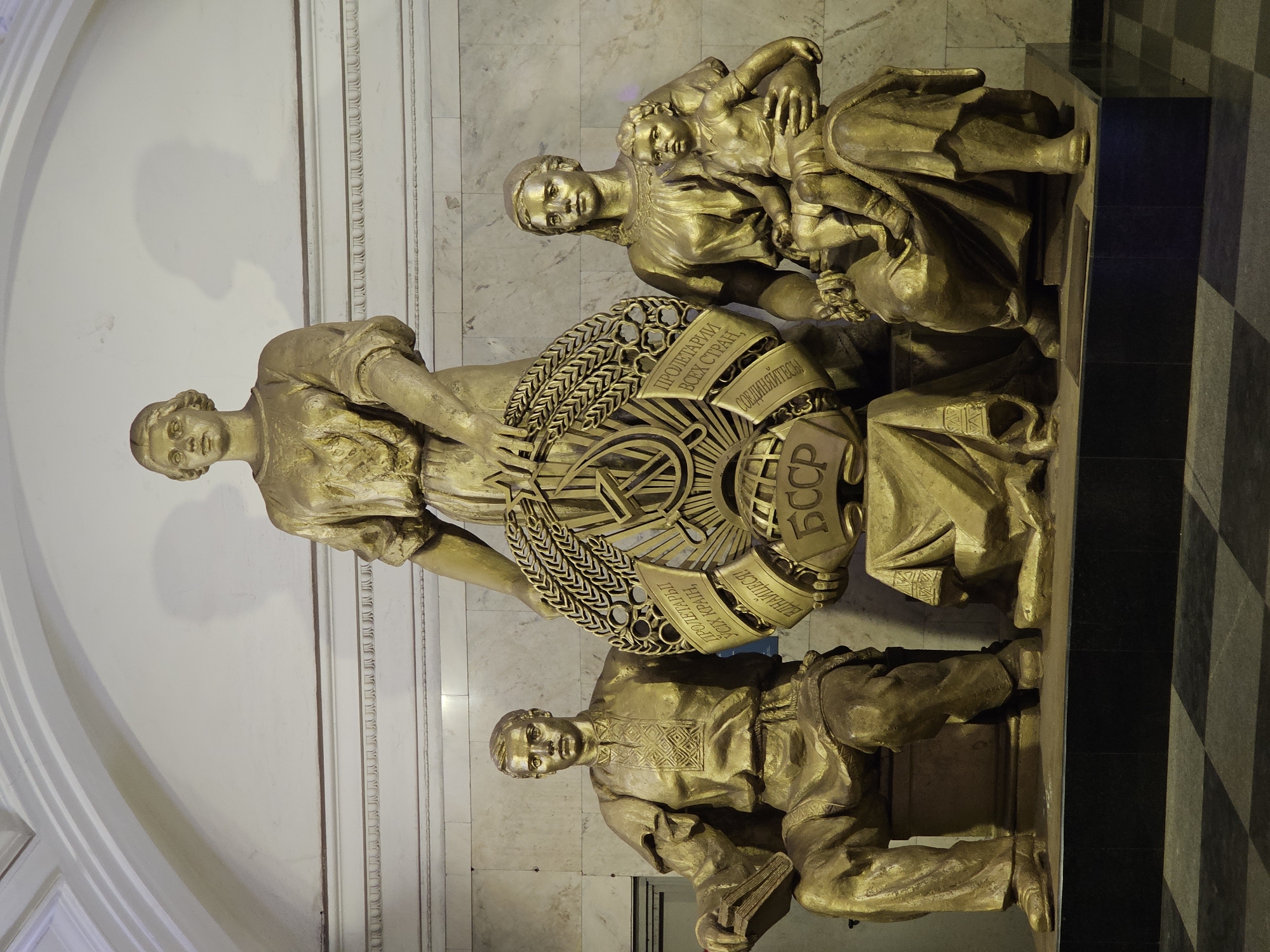 The image shows a bronze sculpture depicting idealized figures of Soviet citizens, likely found in the Moscow Metro, Russia. A female figure stands at the center, holding a wreath around the coat of arms of the USSR. A mother and child are positioned above the female figure. A male figure sits below her, holding a book. The sculpture stands against a wall of white marble with dark stripes on the side.